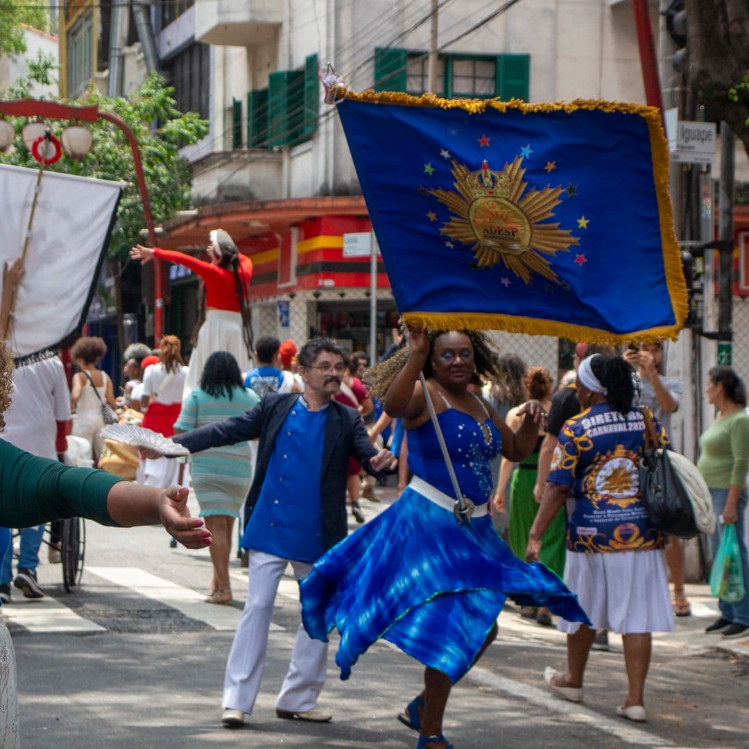 Em cortejo de rua, está dançando um casal de mestre-sala e porta bandeira, composto por uma mulher negra de cabelo cacheado solto, vestindo vestido azul com detalhes brancos e pedrarias e um homem pardo, vestindo terno azul com calça branca, que está segurando um leque branco aberto. A mulher está carregando bandeira azul com franjas e desenho dourados escrito "ADESP". Ao fundo e ao redor, pessoas seguindo o cortejo, árvore, edificações, lanternas "oriental" e postes com fiação elétrica.