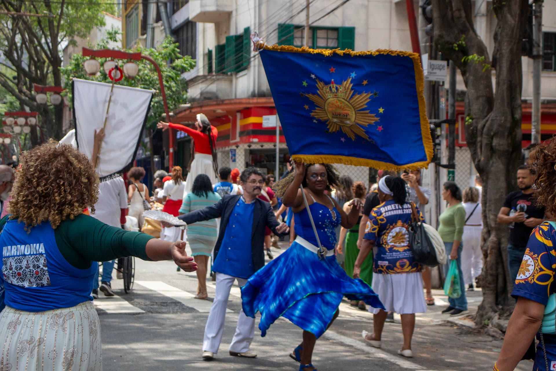 Em cortejo de rua, está dançando um casal de mestre-sala e porta bandeira, composto por uma mulher negra de cabelo cacheado solto, vestindo vestido azul com detalhes brancos e pedrarias e um homem pardo, vestindo terno azul com calça branca, que está segurando um leque branco aberto. A mulher está carregando bandeira azul com franjas e desenho dourados escrito "ADESP". Ao fundo e ao redor, pessoas seguindo o cortejo, árvore, edificações, lanternas "oriental" e postes com fiação elétrica.