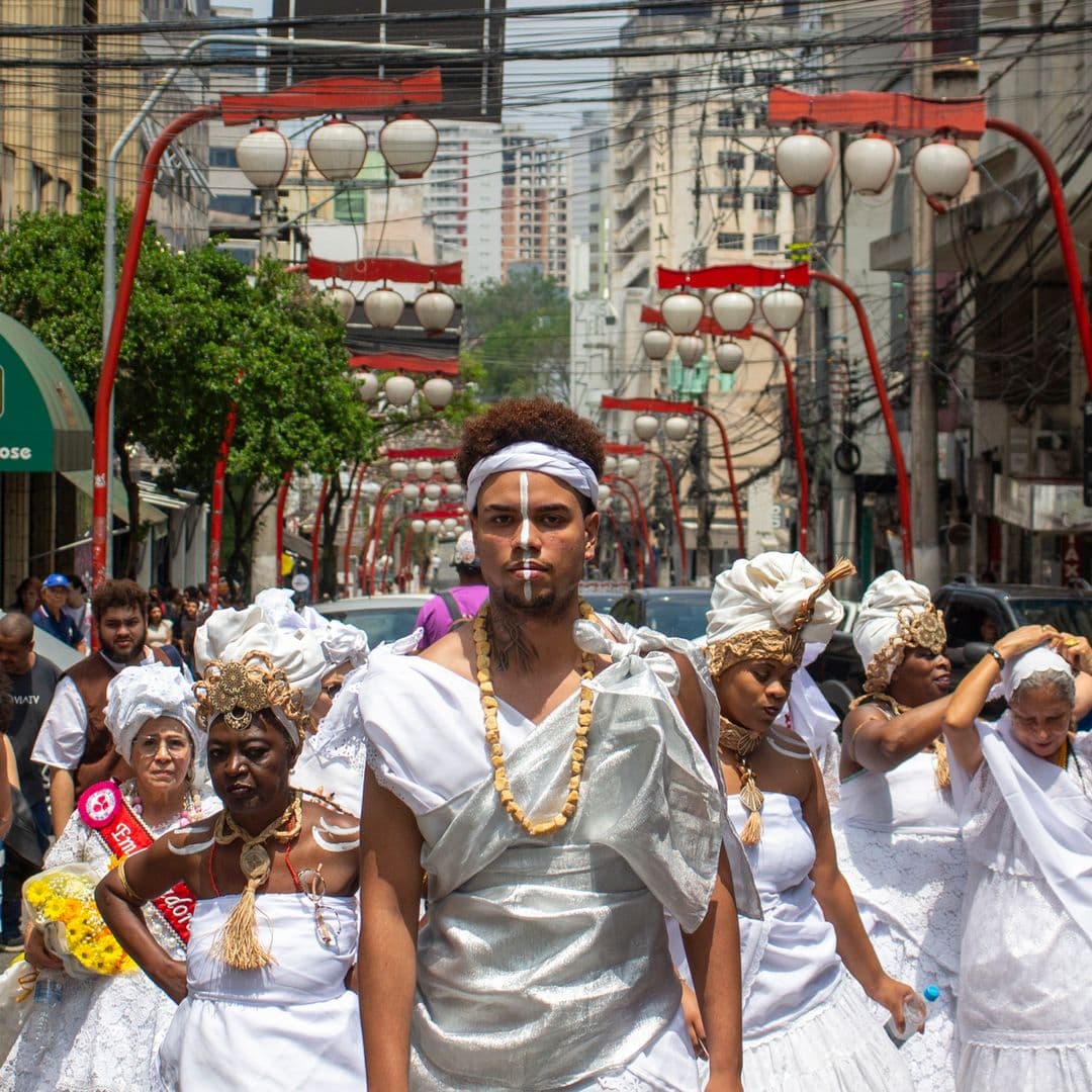 Em cortejo de rua, jovem negro no centro e mulheres vestidas de baiana, todos estão vestidos de branco com elementos simbólicos de matriz africana, como, turbantes de cabeça, saias rodadas, tecidos rendados, adornos, pinturas corporais, e, acessórios artesanais de produtos naturais como a palha, além de uma das baianas a esquerda está com faixa vermelha sob o peitoral escrito "{Embaixadora do Samba}" e nas mãos um buquê de flores na tonalidade amarela, e à direita da imagem, uma mulher está segurando a imagem de Nossa Senhora de Aparecida. Ao fundo e ao redor, pessoas seguindo o cortejo, edificações, árvores, lanternas "oriental", automóveis, placas de sinalização e postes com fiação elétrica.