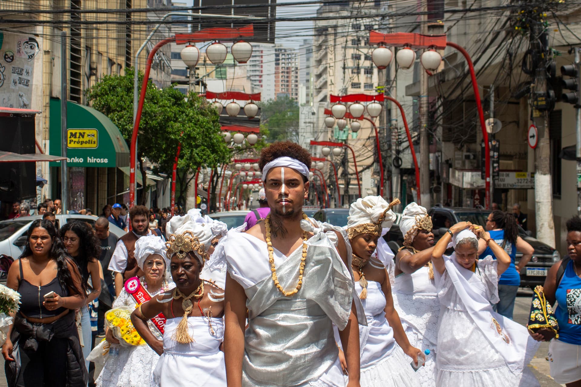 Em cortejo de rua, jovem negro no centro e mulheres vestidas de baiana, todos estão vestidos de branco com elementos simbólicos de matriz africana, como, turbantes de cabeça, saias rodadas, tecidos rendados, adornos, pinturas corporais, e, acessórios artesanais de produtos naturais como a palha, além de uma das baianas a esquerda está com faixa vermelha sob o peitoral escrito "{Embaixadora do Samba}" e nas mãos um buquê de flores na tonalidade amarela, e à direita da imagem, uma mulher está segurando a imagem de Nossa Senhora de Aparecida. Ao fundo e ao redor, pessoas seguindo o cortejo, edificações, árvores, lanternas "oriental", automóveis, placas de sinalização e postes com fiação elétrica.