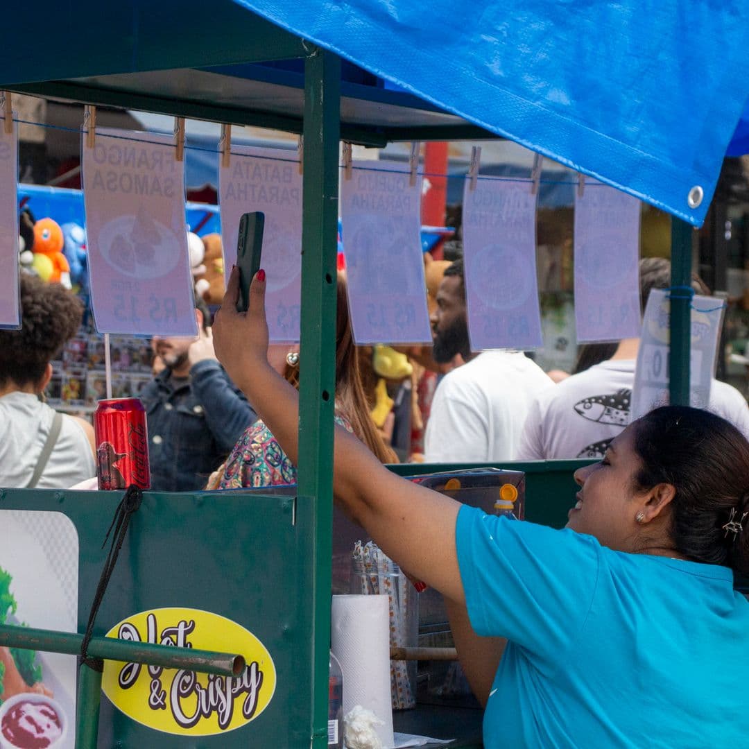 Mulher de cabelo preso e camiseta azul dentro de barraca de venda de comida segura celular registrando o cortejo passando. Ao fundo diversas pessoas pela rua.