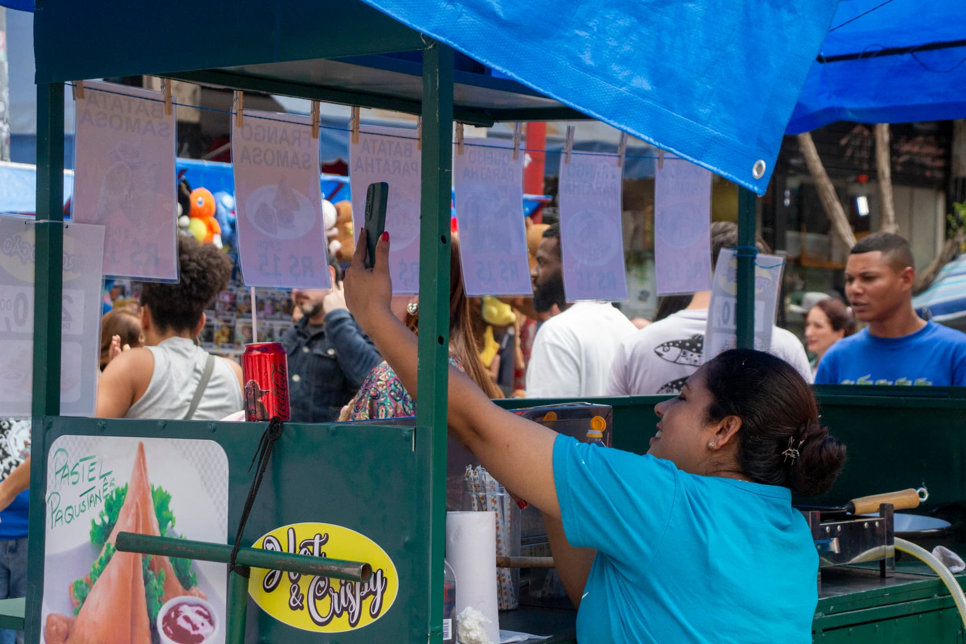 Mulher de cabelo preso e camiseta azul dentro de barraca de venda de comida segura celular registrando o cortejo passando. Ao fundo diversas pessoas pela rua.