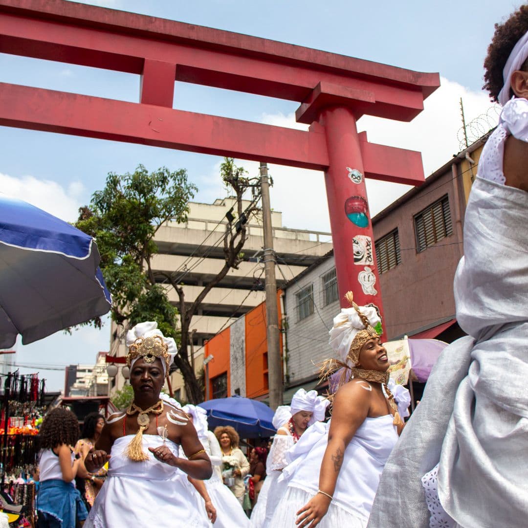 Em cortejo de rua, mulheres negras vestidas de baianas. Estão vestidas de branco com elementos simbólicos de matriz africana, como, turbantes de cabeça, saias rodadas, adornos, pinturas corporais e acessórios artesanais de produtos naturais como a palha. Ao fundo, pessoas seguindo o cortejo, barracas de venda, árvore, edificações, e arco "oriental".