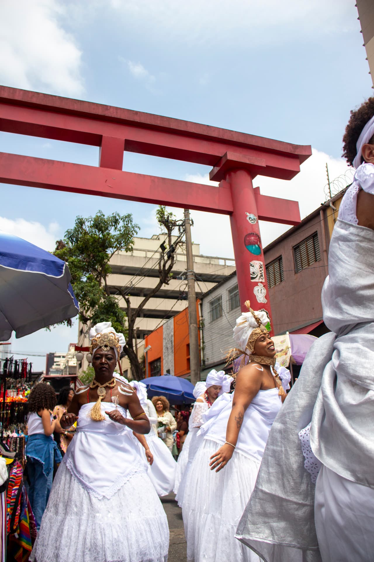 Em cortejo de rua, mulheres negras vestidas de baianas. Estão vestidas de branco com elementos simbólicos de matriz africana, como, turbantes de cabeça, saias rodadas, adornos, pinturas corporais e acessórios artesanais de produtos naturais como a palha. Ao fundo, pessoas seguindo o cortejo, barracas de venda, árvore, edificações, e arco "oriental".