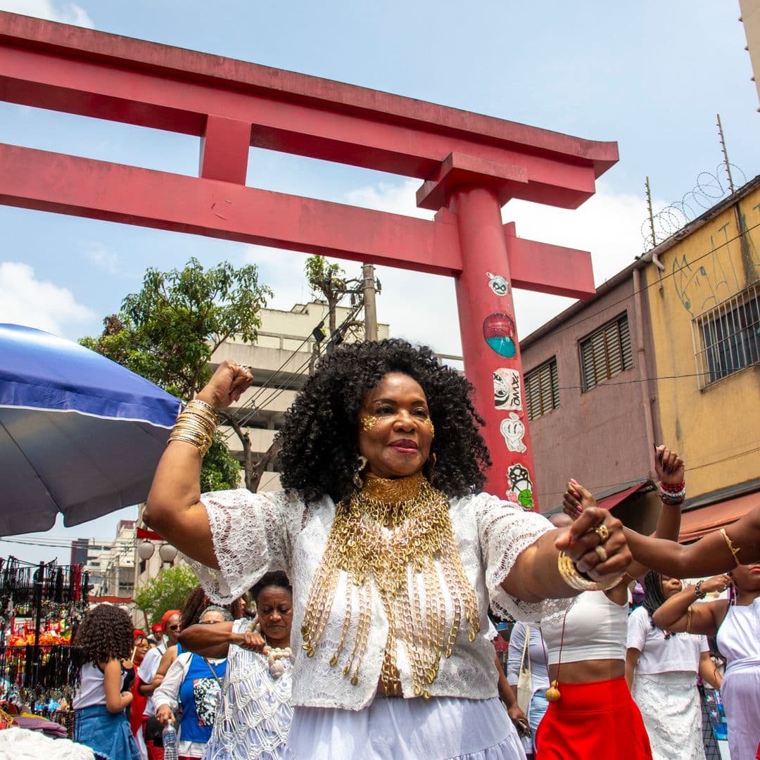 Em cortejo de rua, mulheres negras dançando com elementos simbólicos, que remetem a entidades de matriz africana, como acessórios dourados e espelho de Oxum, cetro branco com pérolas e conchas na mão e colar de conchas de caramujo com esferas grandes bege e branca de Oxalá.  Ao fundo, pessoas seguindo o cortejo, barracas de venda, árvore, edificações, e arco "oriental".