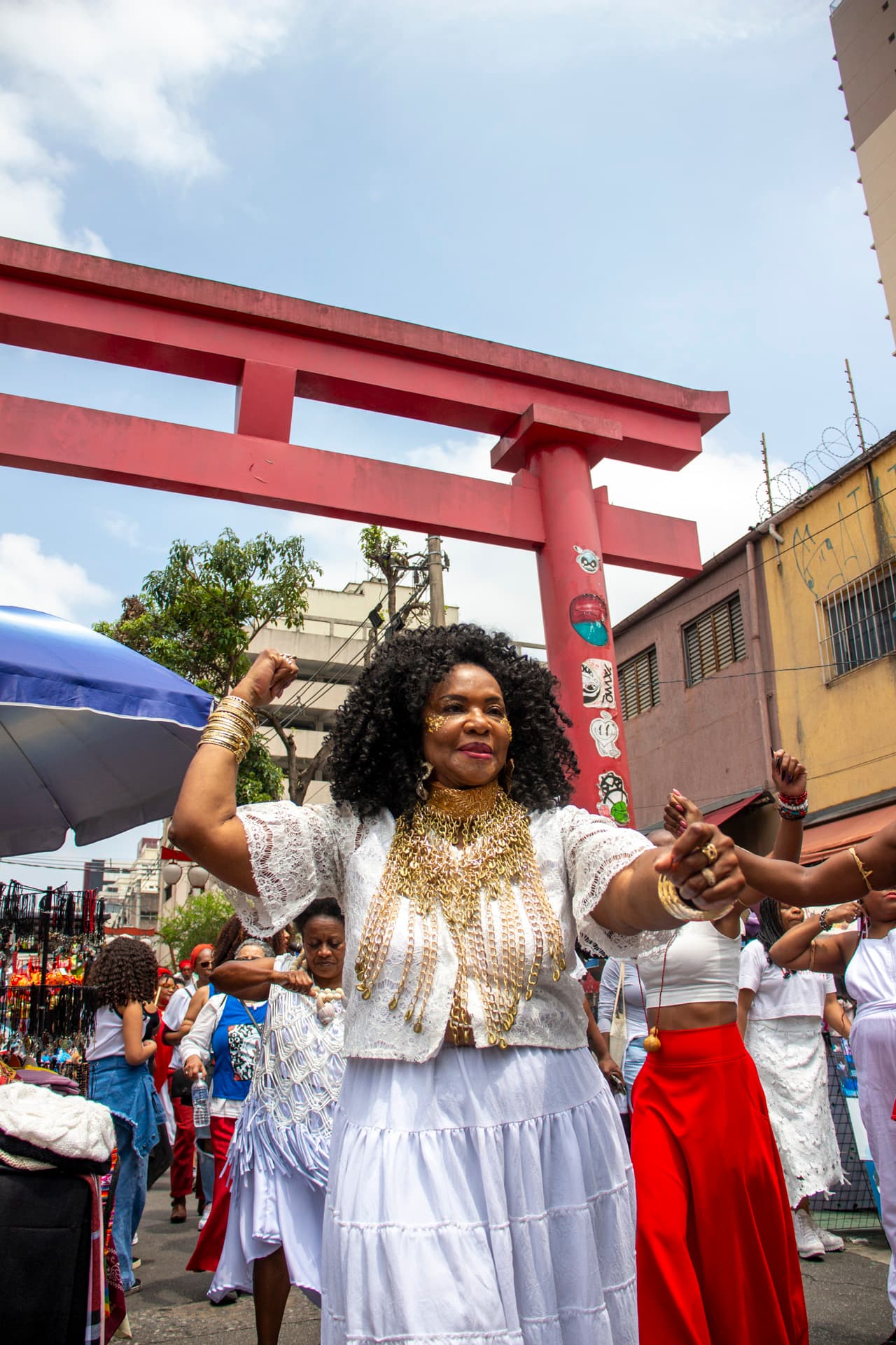Em cortejo de rua, mulheres negras dançando com elementos simbólicos, que remetem a entidades de matriz africana, como acessórios dourados e espelho de Oxum, cetro branco com pérolas e conchas na mão e colar de conchas de caramujo com esferas grandes bege e branca de Oxalá.  Ao fundo, pessoas seguindo o cortejo, barracas de venda, árvore, edificações, e arco "oriental".