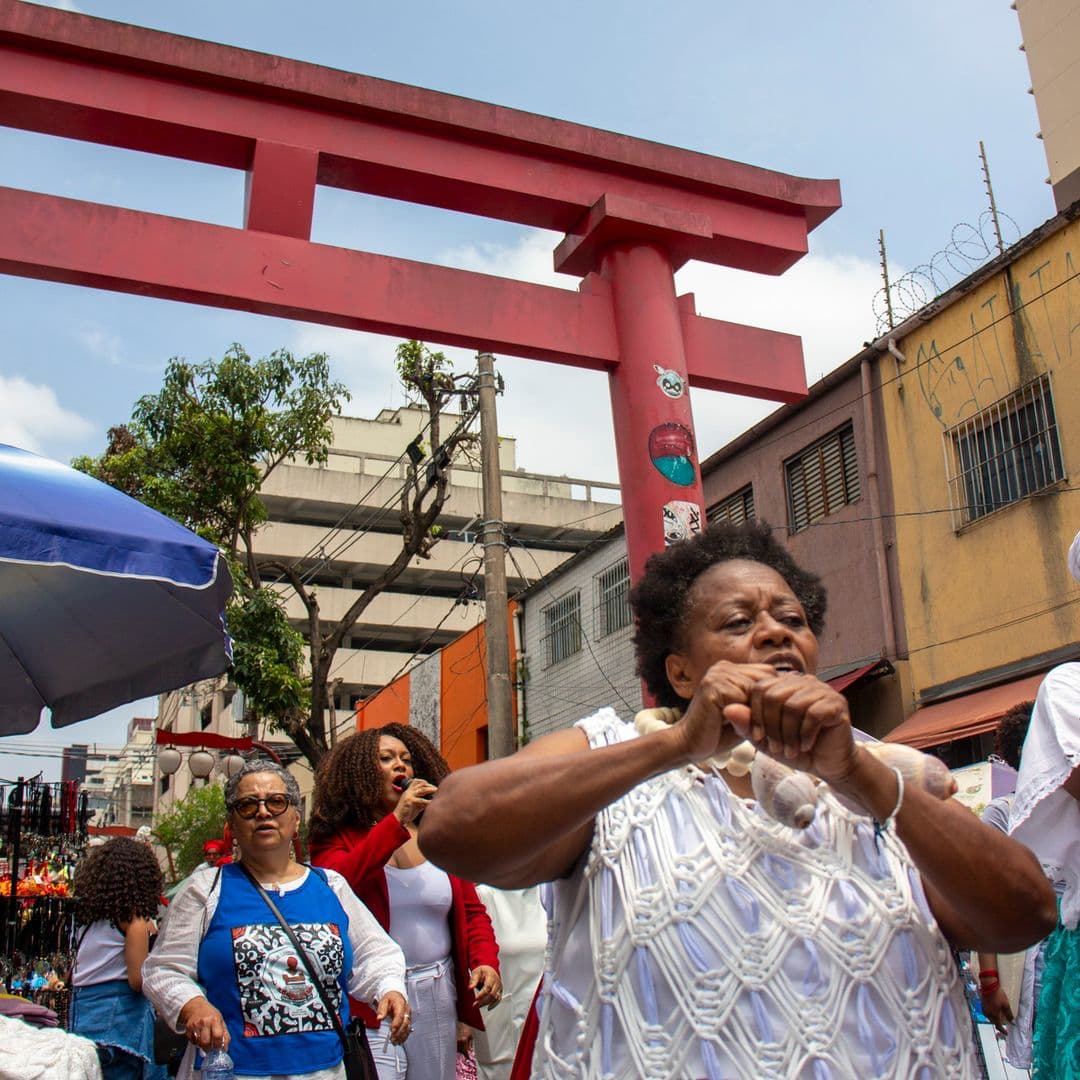 Em cortejo de rua, mulheres negras cantando e dançando com elementos simbólicos, que remetem a entidades de matriz africana, como colar de conchas de caramujo com esferas grandes bege e branca de Oxalá. Ao fundo, mulher vestida de branco e vermelho com microfone na mão cantando, pessoas seguindo o cortejo, barracas de venda, árvore, edificações, e arco "oriental".