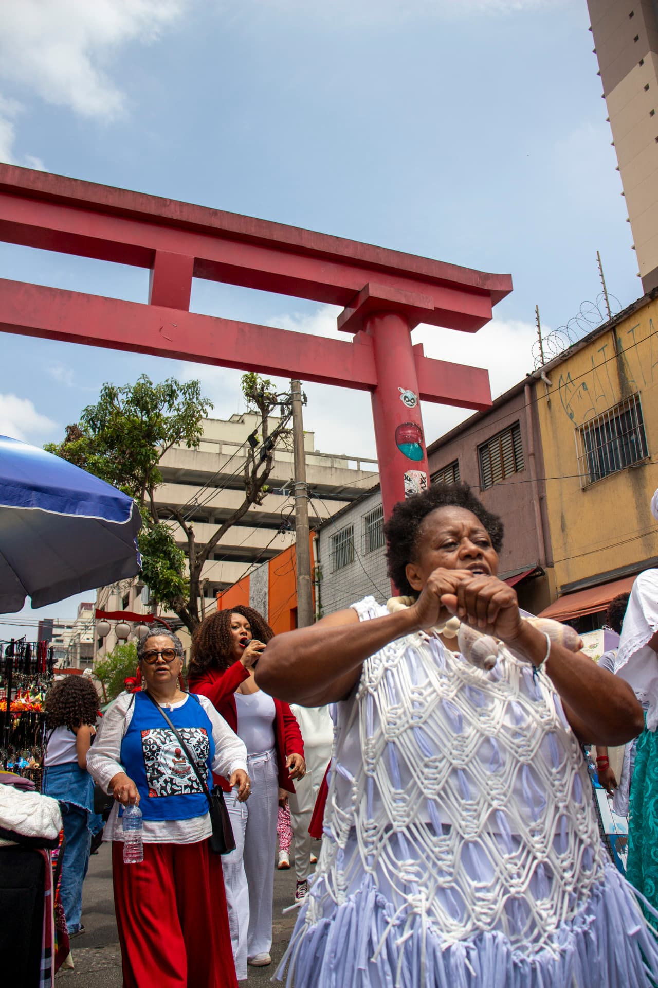 Em cortejo de rua, mulheres negras cantando e dançando com elementos simbólicos, que remetem a entidades de matriz africana, como colar de conchas de caramujo com esferas grandes bege e branca de Oxalá. Ao fundo, mulher vestida de branco e vermelho com microfone na mão cantando, pessoas seguindo o cortejo, barracas de venda, árvore, edificações, e arco "oriental".