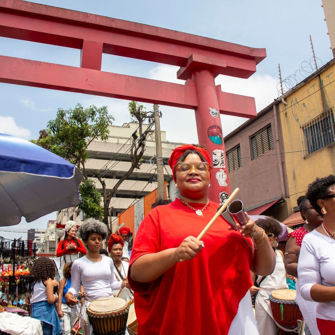 Em cortejo de rua, mulheres negras tocando diversos instrumentos de percussão, estão vestidas majoritariamente de branco e vermelho, acessórios e elementos simbólicos de matriz africana. Ao fundo, duas mulheres negras em elevação ao chão pela utilização de pernas de pau durante apresentação, pessoas seguindo o cortejo, barracas de venda, edificações, arco "oriental, árvores e postes com fiação elétrica.