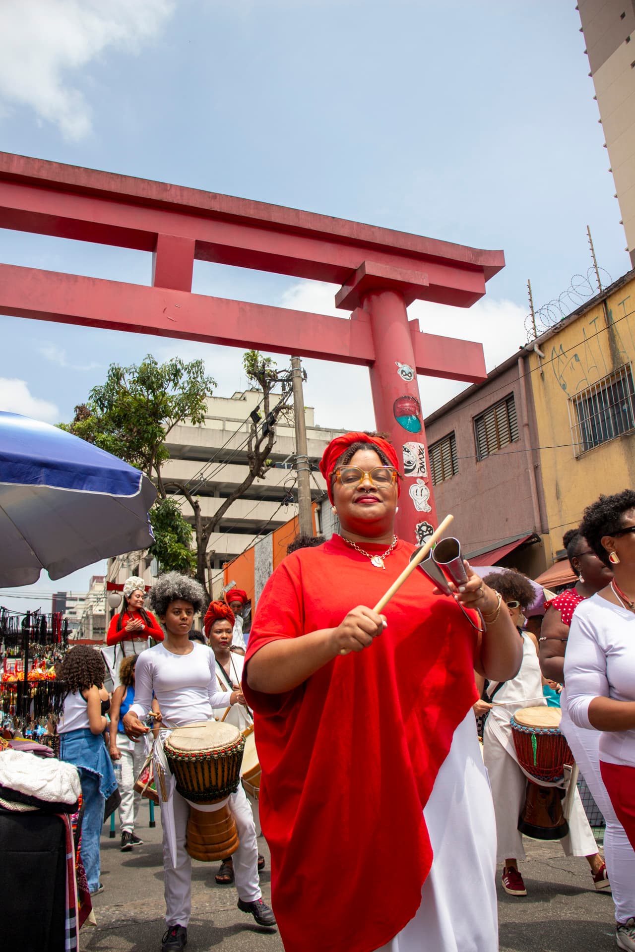 Em cortejo de rua, mulheres negras tocando diversos instrumentos de percussão, estão vestidas majoritariamente de branco e vermelho, acessórios e elementos simbólicos de matriz africana. Ao fundo, duas mulheres negras em elevação ao chão pela utilização de pernas de pau durante apresentação, pessoas seguindo o cortejo, barracas de venda, edificações, arco "oriental, árvores e postes com fiação elétrica.