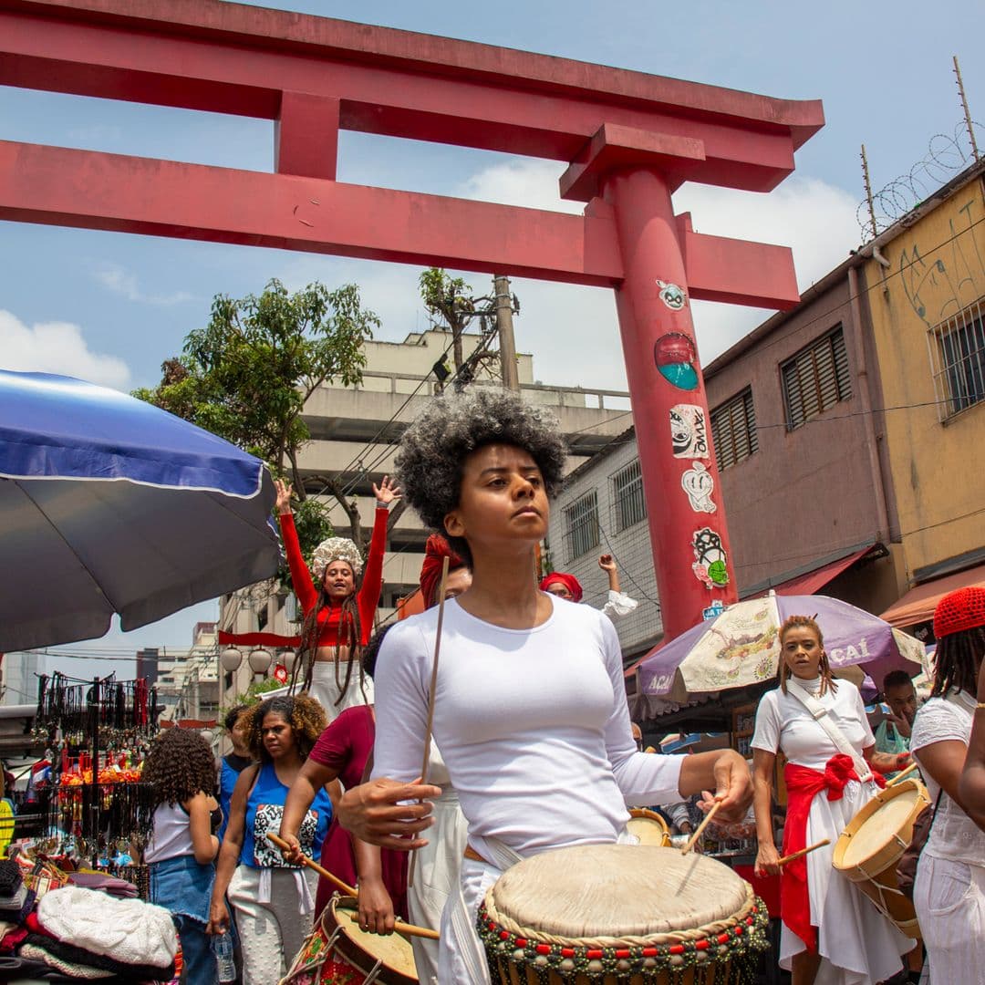 Em cortejo de rua, mulheres negras tocando diversos instrumentos de percussão, estão vestidas majoritariamente de branco e vermelho, acessórios e elementos simbólicos de matriz africana. Ao fundo, duas mulheres negras em elevação ao chão pela utilização de pernas de pau durante apresentação, pessoas seguindo o cortejo, barracas de venda, edificações, arco "oriental, árvores e postes com fiação elétrica.