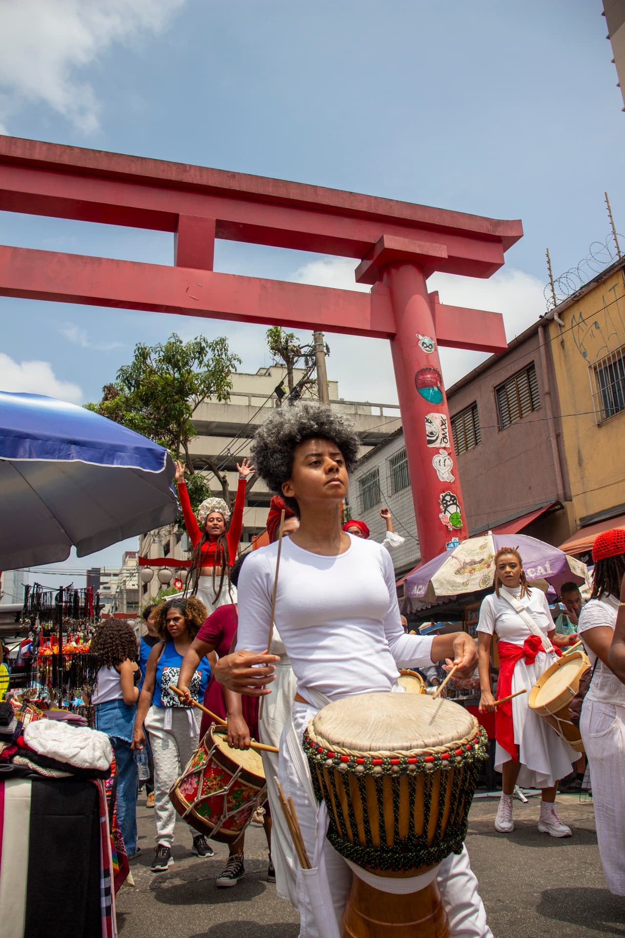 Em cortejo de rua, mulheres negras tocando diversos instrumentos de percussão, estão vestidas majoritariamente de branco e vermelho, acessórios e elementos simbólicos de matriz africana. Ao fundo, duas mulheres negras em elevação ao chão pela utilização de pernas de pau durante apresentação, pessoas seguindo o cortejo, barracas de venda, edificações, arco "oriental, árvores e postes com fiação elétrica.
