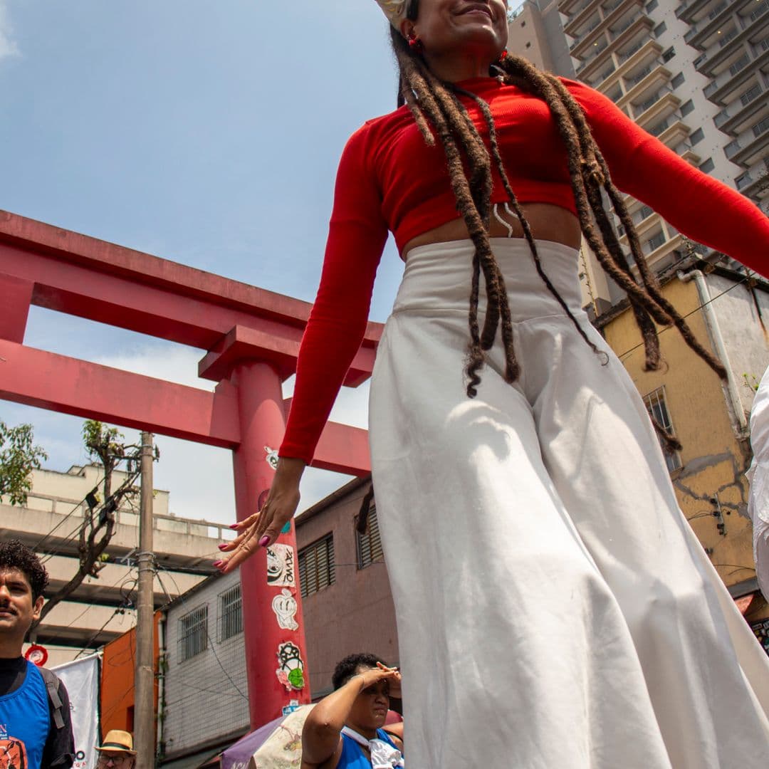 Em cortejo de rua, mulher negra em elevação ao chão pela utilização de pernas de pau durante apresentação vestida de branco e vermelho com acessórios e elementos simbólicos de matriz africana. Ao fundo, pessoas seguindo o cortejo, barracas de venda, edificações, arco "oriental, árvores e postes com fiação elétrica.