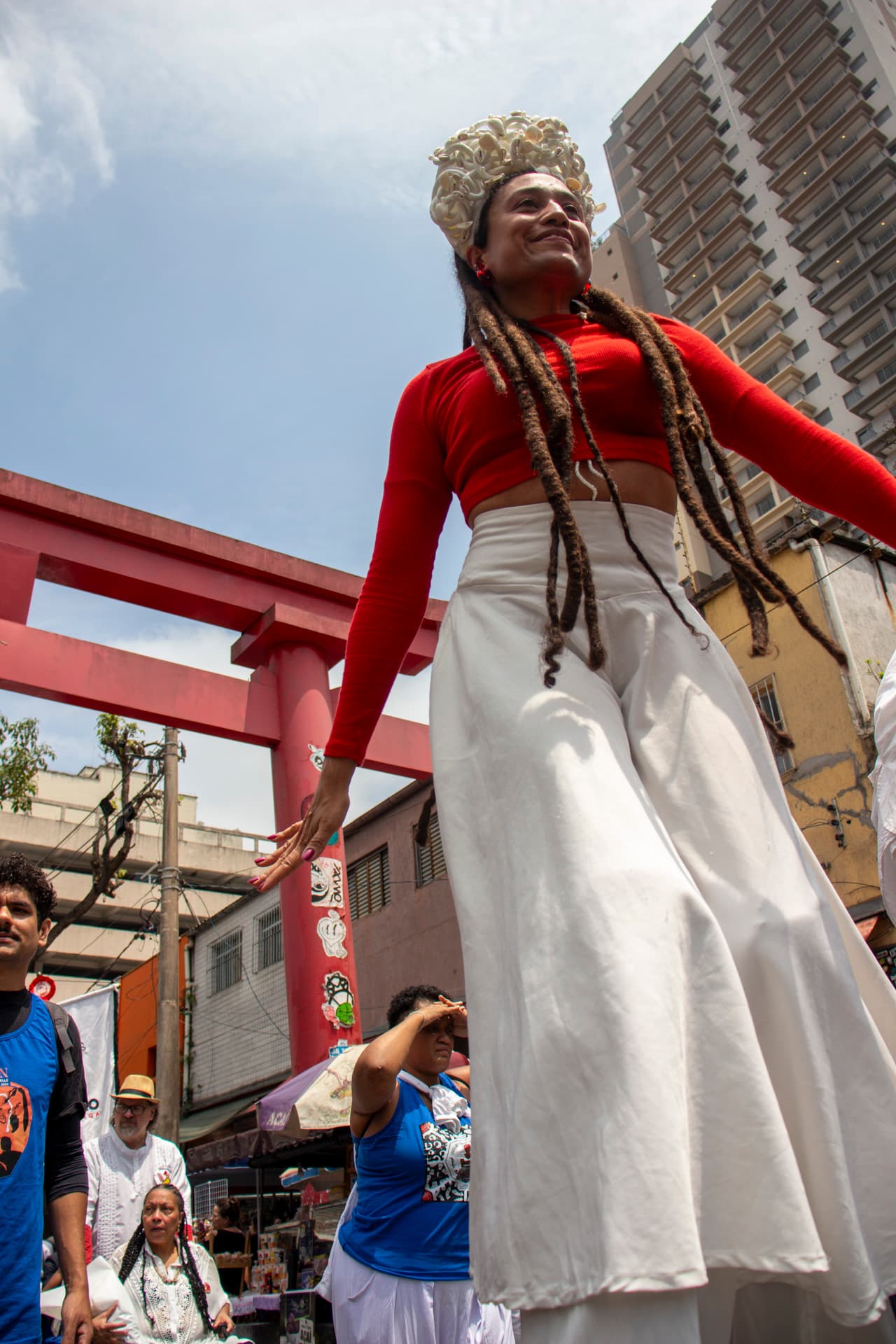 Em cortejo de rua, mulher negra em elevação ao chão pela utilização de pernas de pau durante apresentação vestida de branco e vermelho com acessórios e elementos simbólicos de matriz africana. Ao fundo, pessoas seguindo o cortejo, barracas de venda, edificações, arco "oriental, árvores e postes com fiação elétrica.