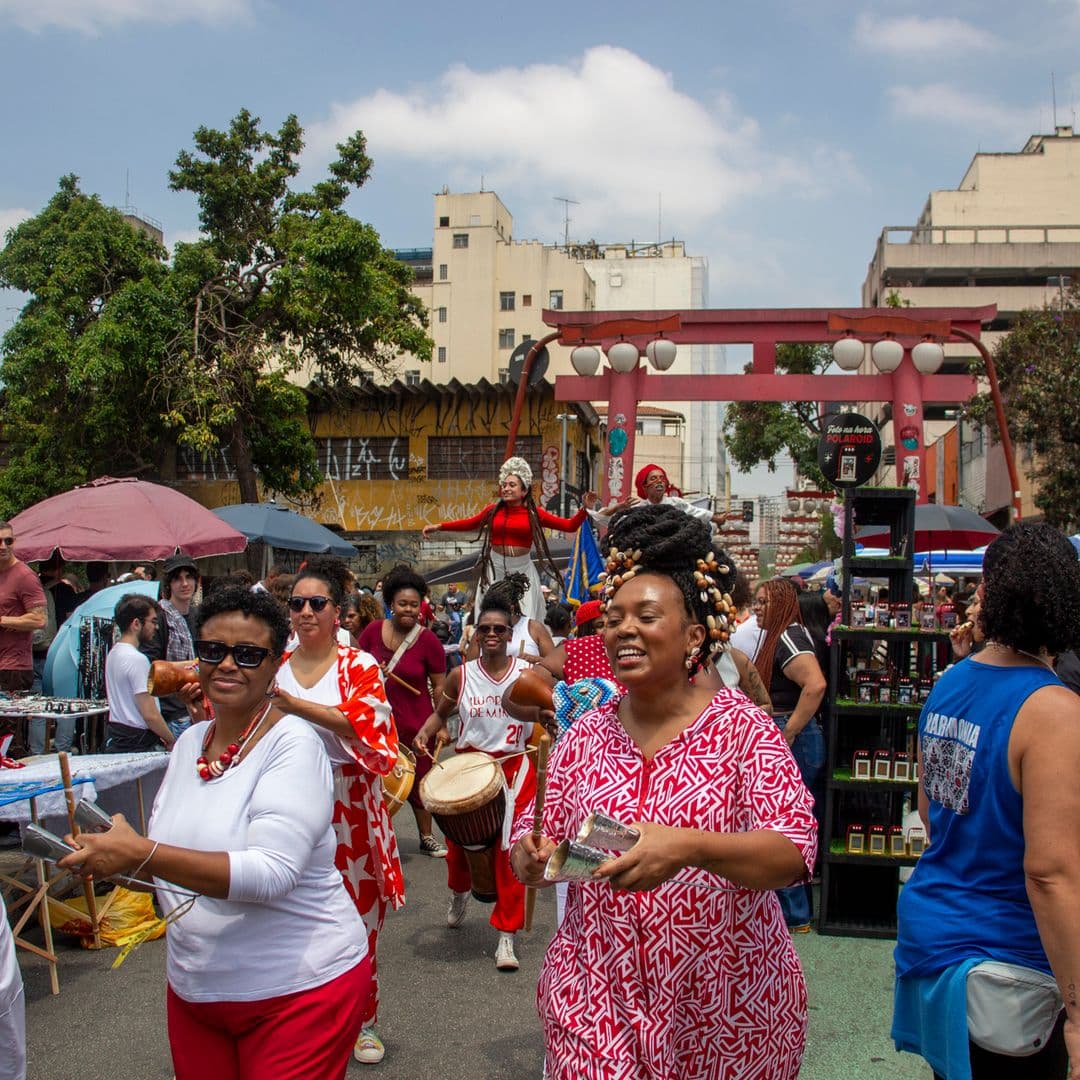 Em cortejo de rua, mulheres negras tocando diversos instrumentos de percussão, estão vestidas majoritariamente de branco e vermelho, algumas com grafismos nas roupas, acessórios e elementos simbólicos de matriz africana. Ao fundo e ao redor, duas mulheres negras em elevação ao chão pela utilização de pernas de pau durante apresentação, pessoas seguindo o cortejo, barracas de venda, edificações, arco e lanternas "oriental e árvores.