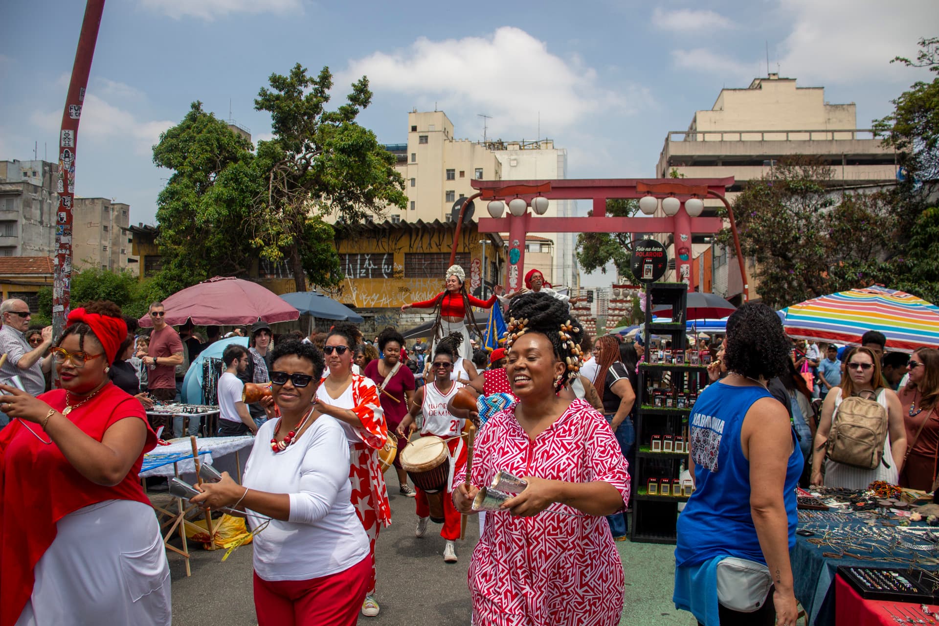 Em cortejo de rua, mulheres negras tocando diversos instrumentos de percussão, estão vestidas majoritariamente de branco e vermelho, algumas com grafismos nas roupas, acessórios e elementos simbólicos de matriz africana. Ao fundo e ao redor, duas mulheres negras em elevação ao chão pela utilização de pernas de pau durante apresentação, pessoas seguindo o cortejo, barracas de venda, edificações, arco e lanternas "oriental e árvores.