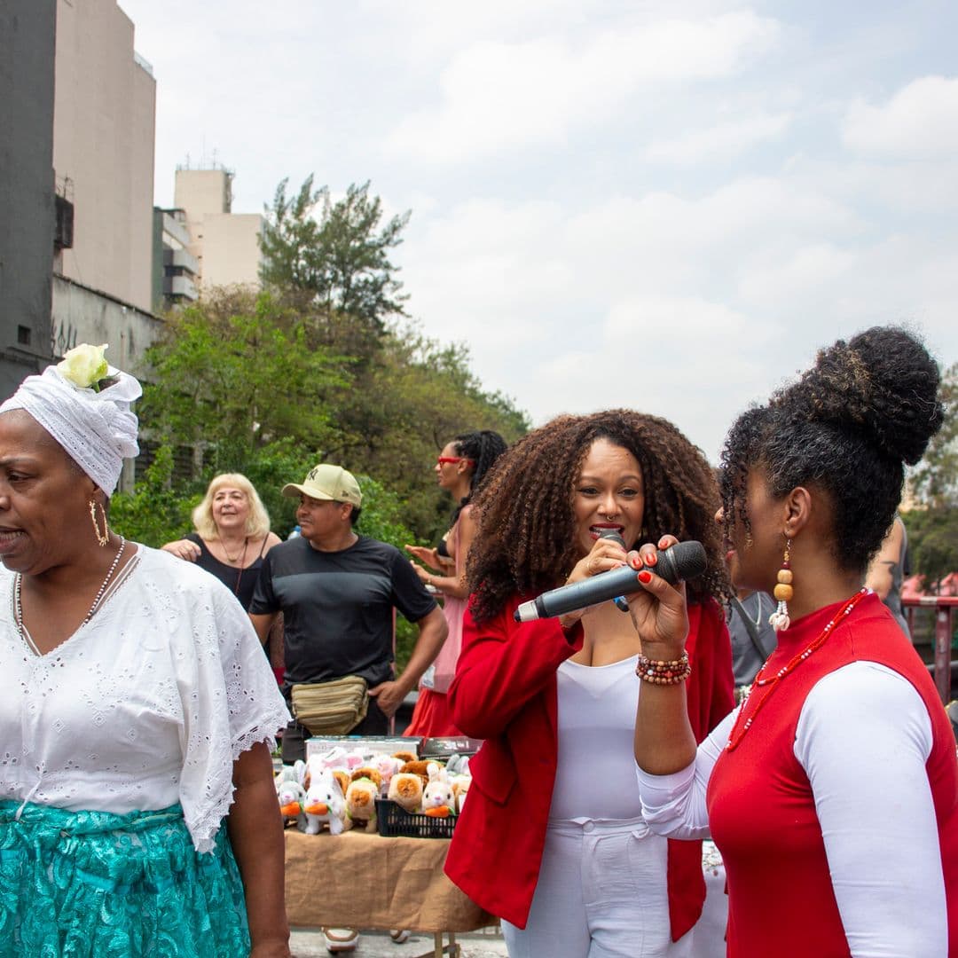 Em cortejo de rua, duas mulheres negras de cabelos cacheados vestidas de vermelho e branco e uma mulher negra de turbante e blusa branca com saia azul, todas com microfones próximo a boca cantando. Ao fundo e ao redor, pessoas seguindo o cortejo, vendedores com barracas de venda, árvores e edificações.