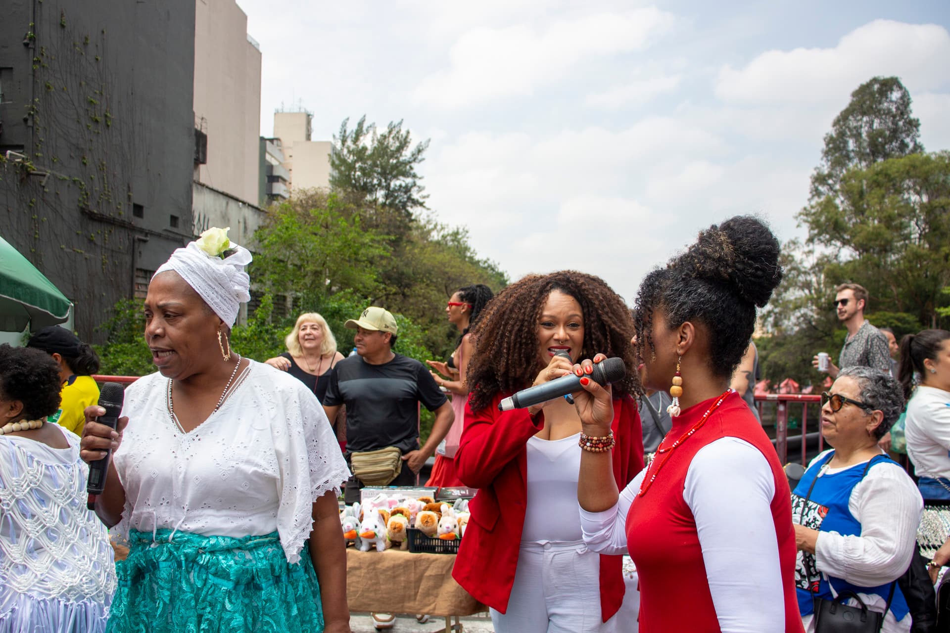 Em cortejo de rua, duas mulheres negras de cabelos cacheados vestidas de vermelho e branco e uma mulher negra de turbante e blusa branca com saia azul, todas com microfones próximo a boca cantando. Ao fundo e ao redor, pessoas seguindo o cortejo, vendedores com barracas de venda, árvores e edificações.