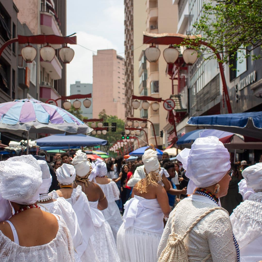 Em cortejo de rua, grupo predominante de mulheres de costas, vestidas majoritariamente de branco com elementos simbólicos de matriz africana, como, turbantes de cabeça, saias rodadas, colar de contas nas cores vermelho, branco e preto, e, acessórios artesanais de produtos naturais como a palha. Ao fundo e ao redor, pessoas assistindo e outras seguindo o cortejo, barracas de venda, edificações, árvore, lanternas "oriental" e postes com fiação elétrica.