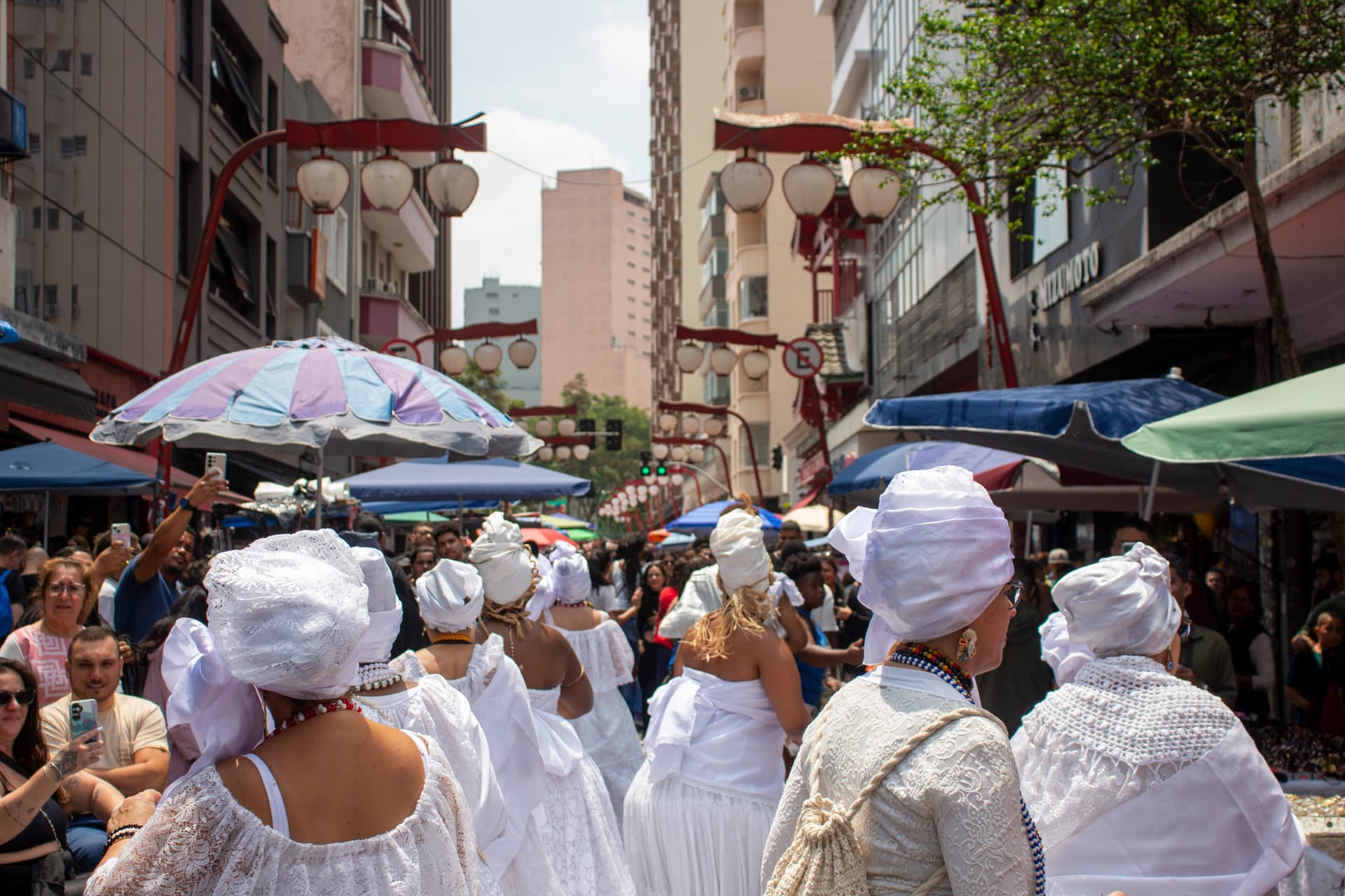 Em cortejo de rua, grupo predominante de mulheres de costas, vestidas majoritariamente de branco com elementos simbólicos de matriz africana, como, turbantes de cabeça, saias rodadas, colar de contas nas cores vermelho, branco e preto, e, acessórios artesanais de produtos naturais como a palha. Ao fundo e ao redor, pessoas assistindo e outras seguindo o cortejo, barracas de venda, edificações, árvore, lanternas "oriental" e postes com fiação elétrica.
