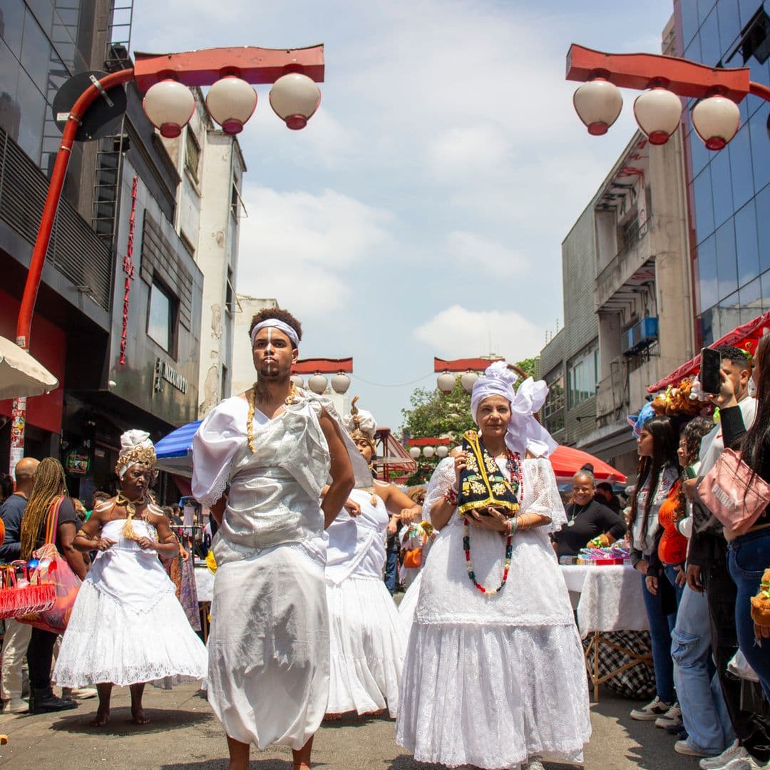 Em cortejo de rua, jovem negro no centro e mulheres vestidas de baiana dançando, todos estão vestidos de branco com elementos simbólicos de matriz africana, como, turbantes de cabeça, saias rodadas, tecidos rendados, adornos, pinturas corporais, e, acessórios artesanais de produtos naturais como a palha, além de uma das mulheres, à direita da imagem, estar segurando a imagem de Nossa Senhora de Aparecida. Ao fundo e ao redor, pessoas assistindo e outras seguindo o cortejo, lanternas "oriental", barracas de venda, edificações, árvores.