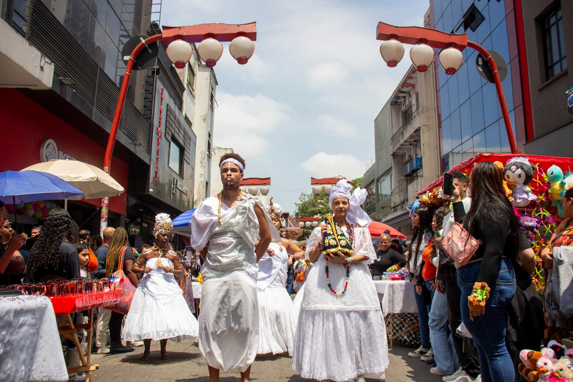 Em cortejo de rua, jovem negro no centro e mulheres vestidas de baiana dançando, todos estão vestidos de branco com elementos simbólicos de matriz africana, como, turbantes de cabeça, saias rodadas, tecidos rendados, adornos, pinturas corporais, e, acessórios artesanais de produtos naturais como a palha, além de uma das mulheres, à direita da imagem, estar segurando a imagem de Nossa Senhora de Aparecida. Ao fundo e ao redor, pessoas assistindo e outras seguindo o cortejo, lanternas "oriental", barracas de venda, edificações, árvores.