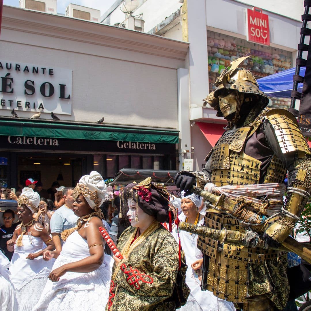 Em cortejo de rua, mulheres vestidas majoritariamente de branco com elementos simbólicos de matriz africana, como, turbantes de cabeça, saias rodadas, tecidos de rendas, e, acessórios artesanais de produtos naturais como a palha. Ao lado, pessoas vestidas de personagens orientais, ao fundo, pessoas assistindo e outras seguindo o cortejo, barracas de venda, edificações e postes com fiação elétrica.