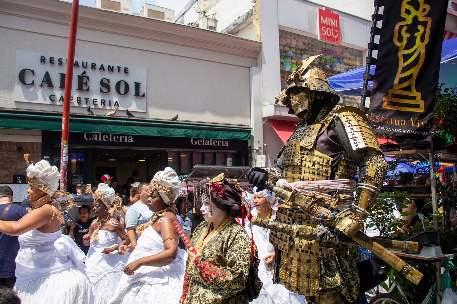 Em cortejo de rua, mulheres vestidas majoritariamente de branco com elementos simbólicos de matriz africana, como, turbantes de cabeça, saias rodadas, tecidos de rendas, e, acessórios artesanais de produtos naturais como a palha. Ao lado, pessoas vestidas de personagens orientais, ao fundo, pessoas assistindo e outras seguindo o cortejo, barracas de venda, edificações e postes com fiação elétrica.
