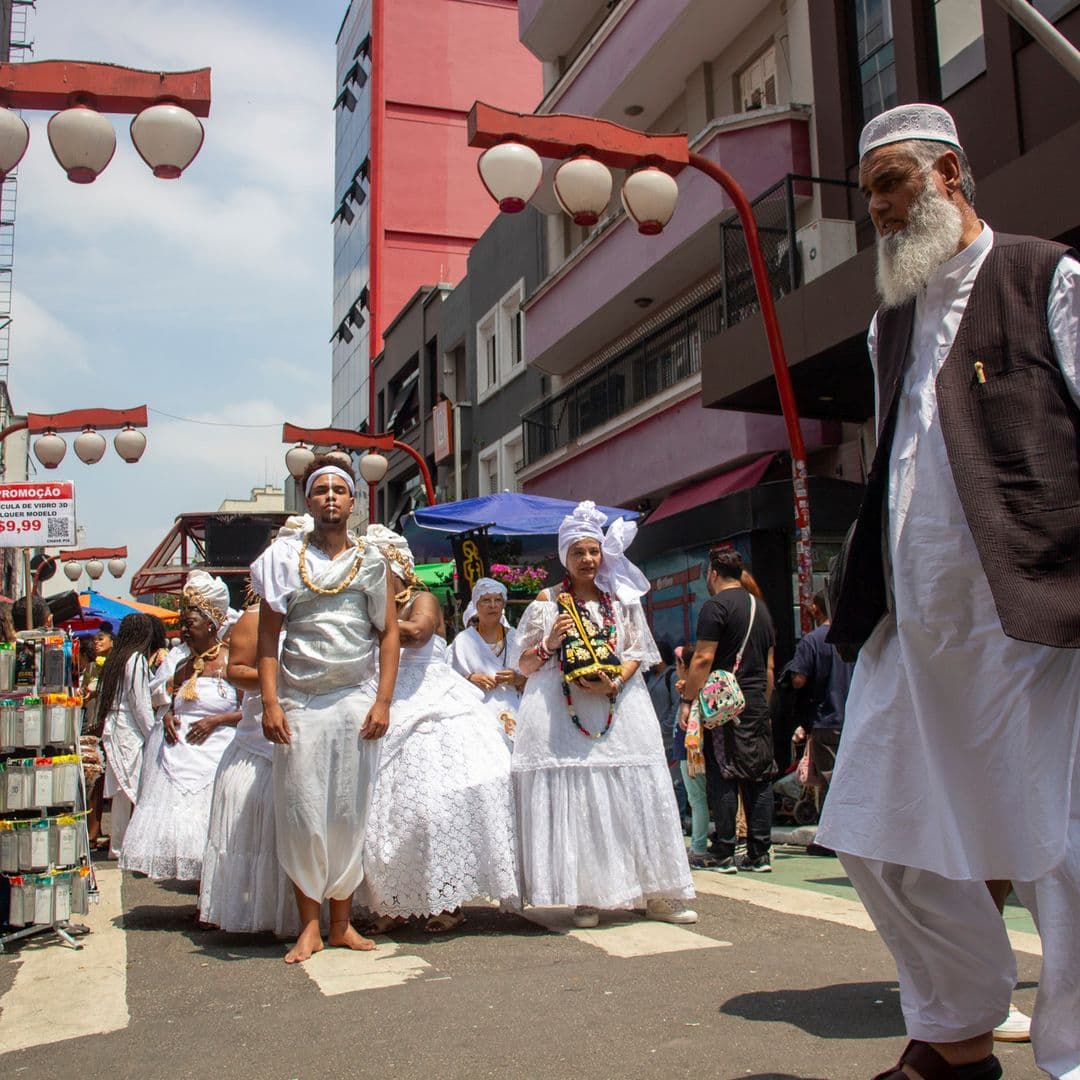 Em cortejo de rua, jovem negro no centro e mulheres vestidas de baiana dançando, todos estão vestidos de branco com elementos simbólicos de matriz africana, como, turbantes de cabeça, saias rodadas, tecidos rendados, adornos, pinturas corporais, e, acessórios artesanais de produtos naturais como a palha, além de uma das mulheres, à direita da imagem, estar segurando a imagem de Nossa Senhora de Aparecida. No canto direito homem com roupas típicas do oriente médio, atravessando a rua. Ao fundo e ao redor, pessoas assistindo e outras seguindo o cortejo, lanternas "oriental", vendedores e barracas de venda e edificações.
