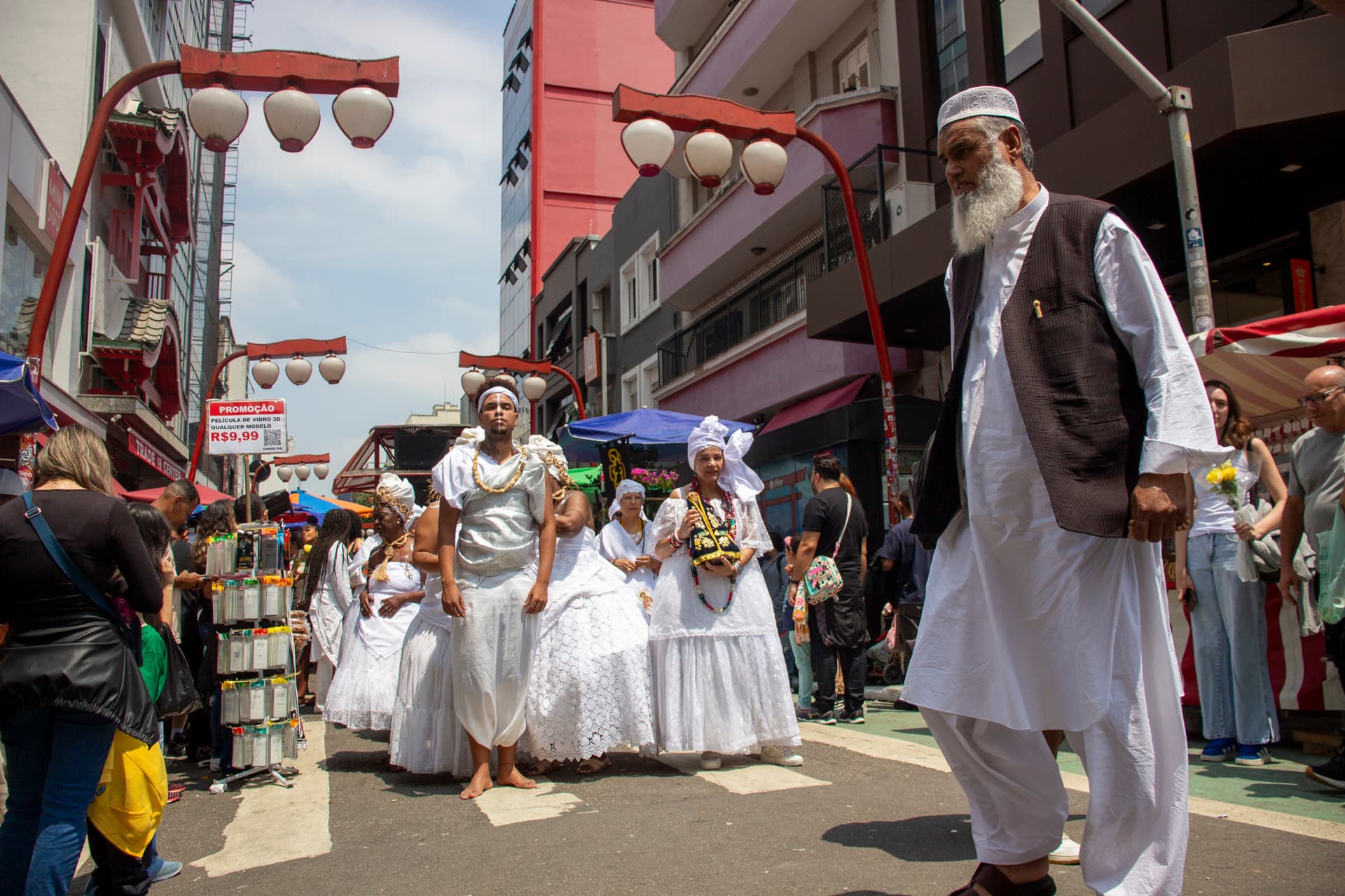 Em cortejo de rua, jovem negro no centro e mulheres vestidas de baiana dançando, todos estão vestidos de branco com elementos simbólicos de matriz africana, como, turbantes de cabeça, saias rodadas, tecidos rendados, adornos, pinturas corporais, e, acessórios artesanais de produtos naturais como a palha, além de uma das mulheres, à direita da imagem, estar segurando a imagem de Nossa Senhora de Aparecida. No canto direito homem com roupas típicas do oriente médio, atravessando a rua. Ao fundo e ao redor, pessoas assistindo e outras seguindo o cortejo, lanternas "oriental", vendedores e barracas de venda e edificações.