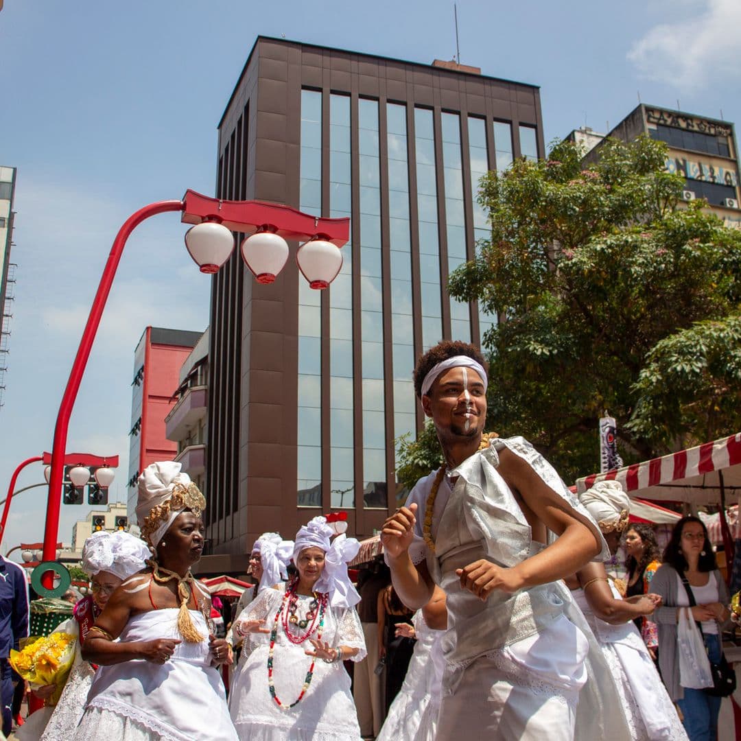 Em cortejo de rua, jovem negro no centro e mulheres vestidas de baiana dançando, todos estão vestidos de branco com elementos simbólicos de matriz africana, como, turbantes de cabeça, saias rodadas, tecidos rendados, adornos, pinturas corporais, e, acessórios artesanais de produtos naturais como a palha, além de uma das mulheres, à direita da imagem, estar segurando a imagem de Nossa Senhora de Aparecida. Ao fundo e ao redor, pessoas assistindo e outras seguindo o cortejo, lanternas "oriental", barracas de venda, edificações e árvores.
