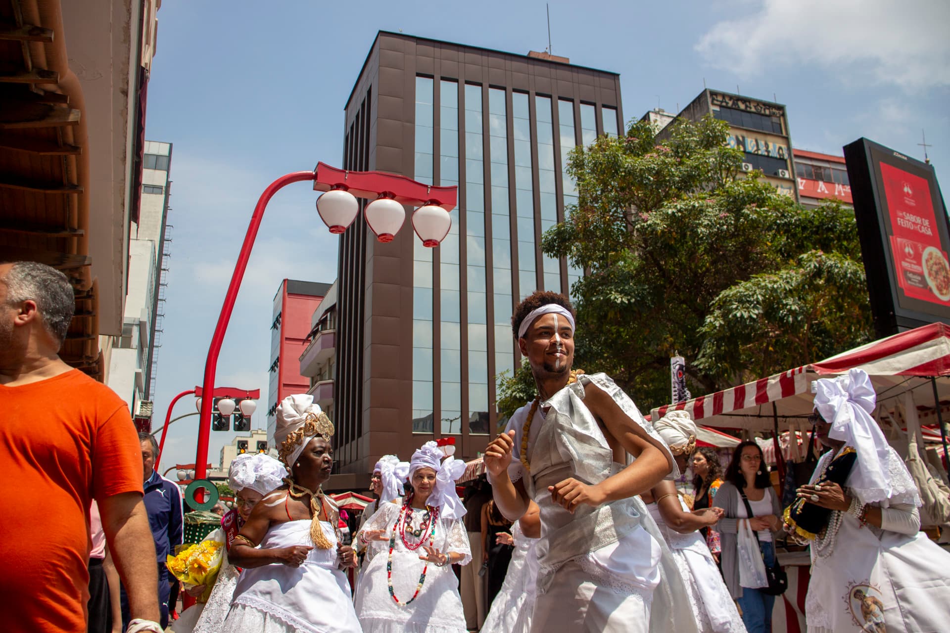 Em cortejo de rua, jovem negro no centro e mulheres vestidas de baiana dançando, todos estão vestidos de branco com elementos simbólicos de matriz africana, como, turbantes de cabeça, saias rodadas, tecidos rendados, adornos, pinturas corporais, e, acessórios artesanais de produtos naturais como a palha, além de uma das mulheres, à direita da imagem, estar segurando a imagem de Nossa Senhora de Aparecida. Ao fundo e ao redor, pessoas assistindo e outras seguindo o cortejo, lanternas "oriental", barracas de venda, edificações e árvores.