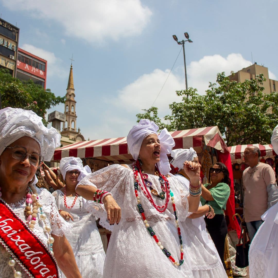 Em cortejo de rua, mulheres vestidas de baianas dançando com roupas brancas e com elementos simbólicos de matriz africana, como, turbantes de cabeça, saias rodadas, tecidos rendados, colar de contas, acessórios artesanais de produtos naturais como a palha, além de uma das mulheres, na lateral esquerda, esta vestindo uma faixa vermelha escrito "Embaixadora {do Samba}" e segurando nas mãos um buquê de flores na tonalidade amarela. Ao fundo, pessoas assistindo e outras seguindo o cortejo, edificações e barracas de venda,.