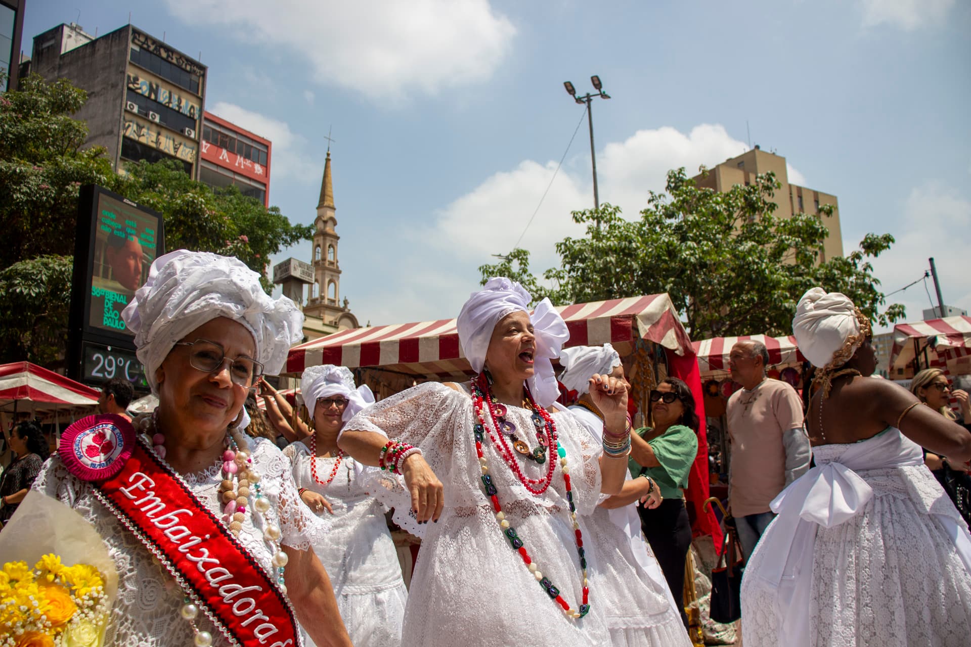 Em cortejo de rua, mulheres vestidas de baianas dançando com roupas brancas e com elementos simbólicos de matriz africana, como, turbantes de cabeça, saias rodadas, tecidos rendados, colar de contas, acessórios artesanais de produtos naturais como a palha, além de uma das mulheres, na lateral esquerda, esta vestindo uma faixa vermelha escrito "Embaixadora {do Samba}" e segurando nas mãos um buquê de flores na tonalidade amarela. Ao fundo, pessoas assistindo e outras seguindo o cortejo, edificações e barracas de venda,.