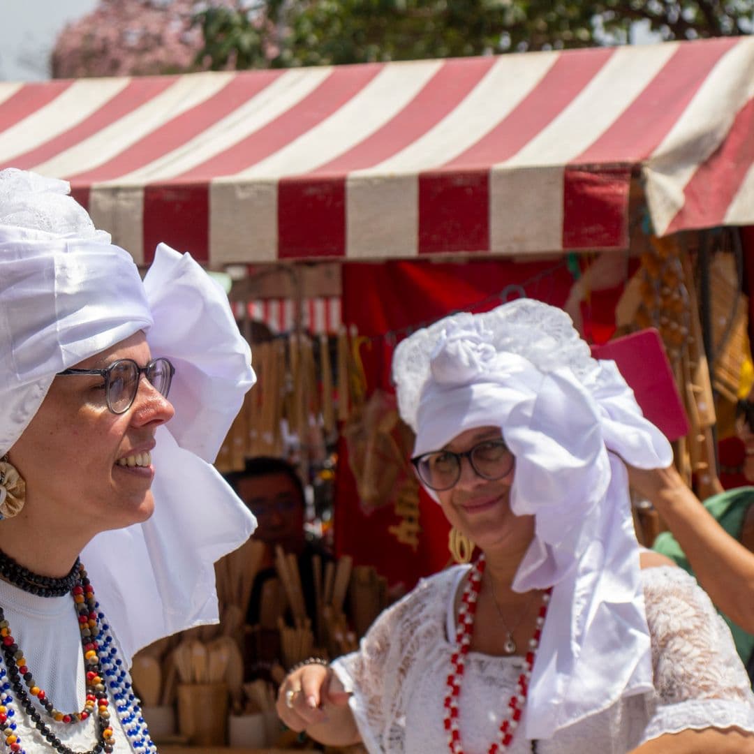 Em cortejo de rua, mulheres vestidas de baianas dançando com roupas brancas e com elementos simbólicos de matriz africana, como, turbantes de cabeça, saias rodadas, tecidos rendados, colar de contas. Ao fundo, pessoas assistindo e outras seguindo o cortejo, barracas de venda e árvore.