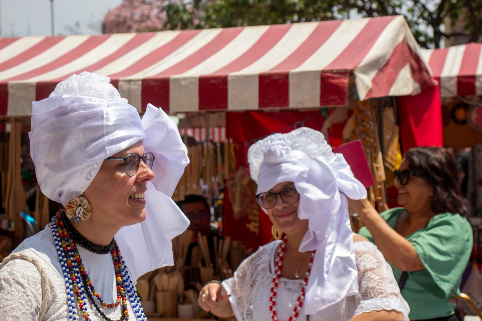 Em cortejo de rua, mulheres vestidas de baianas dançando com roupas brancas e com elementos simbólicos de matriz africana, como, turbantes de cabeça, saias rodadas, tecidos rendados, colar de contas. Ao fundo, pessoas assistindo e outras seguindo o cortejo, barracas de venda e árvore.