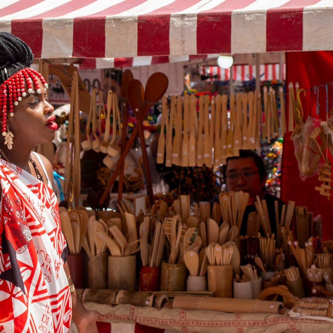 Em cortejo de rua, mulher negra de lado andando, vestida de branco e vermelho com grafismos e elementos simbólicos de matriz africana. Ao fundo, na cor vermelha e branca uma barraca de venda de utensílios de cozinha feitos de bambu com vendedor atrás assistindo o cortejo.