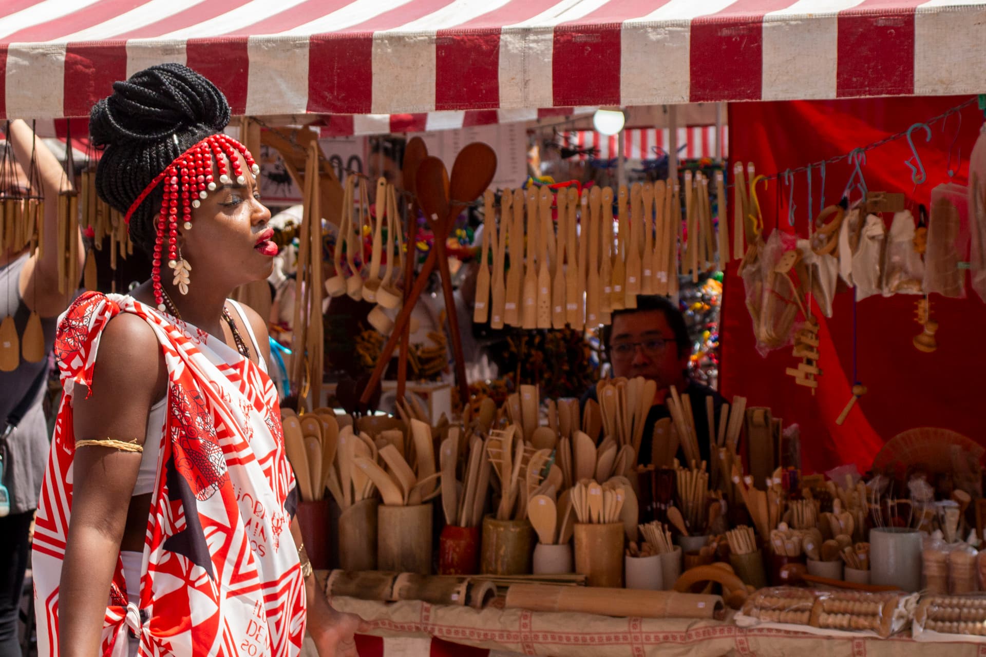 Em cortejo de rua, mulher negra de lado andando, vestida de branco e vermelho com grafismos e elementos simbólicos de matriz africana. Ao fundo, na cor vermelha e branca uma barraca de venda de utensílios de cozinha feitos de bambu com vendedor atrás assistindo o cortejo.