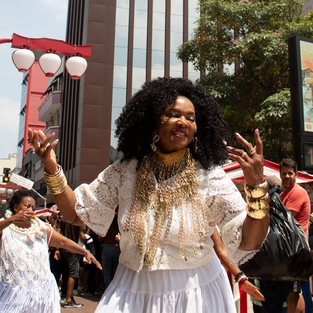 Em cortejo de rua, mulheres negras dançando com elementos simbólicos, que remetem a entidades de matriz africana, como acessórios dourados e espelho de Oxum, colar de conchas de caramujo com esferas grandes bege e branca de Oxalá. Ao fundo e ao redor, pessoas assistindo e seguindo o cortejo, barracas de venda, árvore, edificações, e lanternas "oriental".