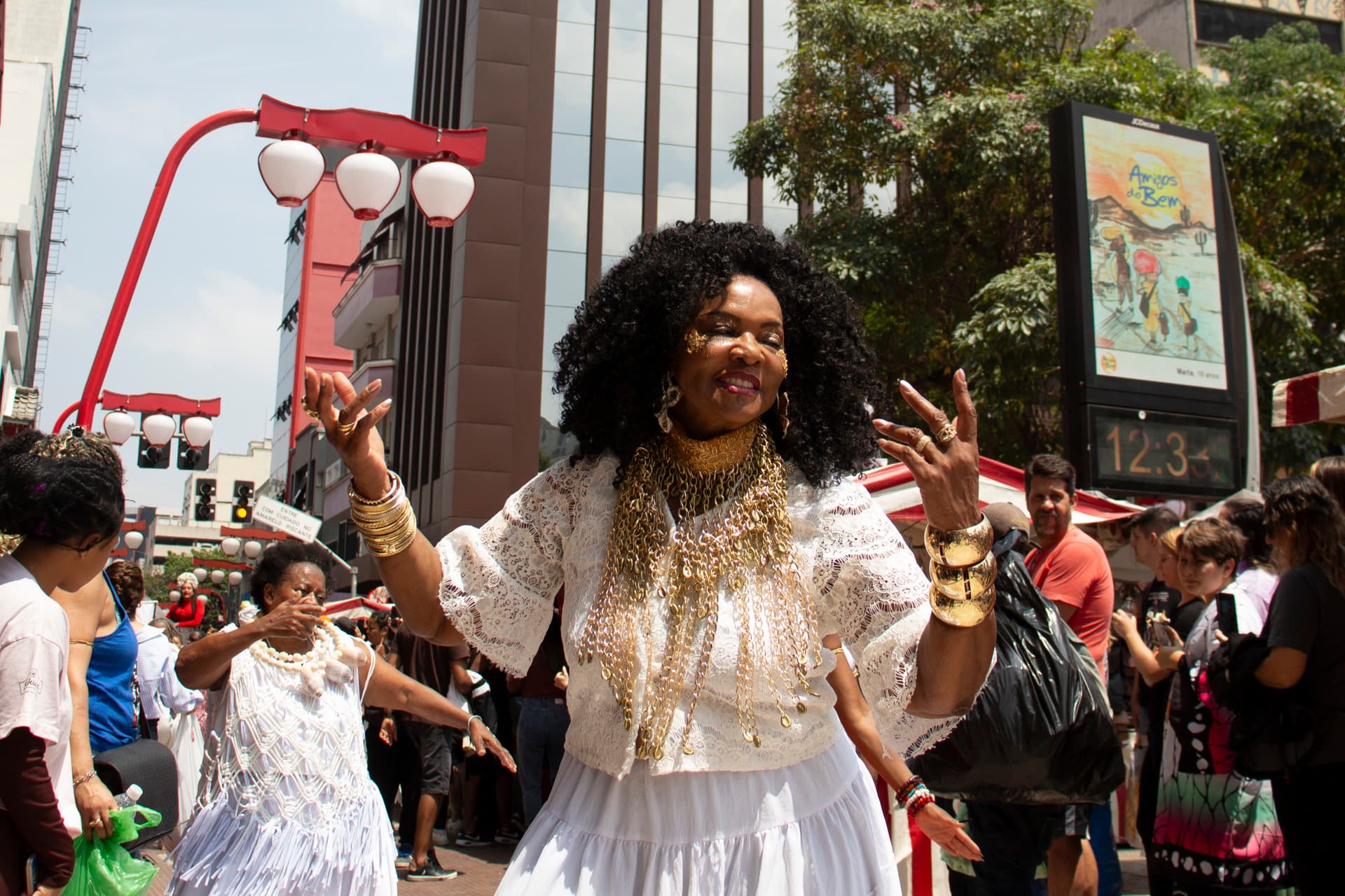 Em cortejo de rua, mulheres negras dançando com elementos simbólicos, que remetem a entidades de matriz africana, como acessórios dourados e espelho de Oxum, colar de conchas de caramujo com esferas grandes bege e branca de Oxalá. Ao fundo e ao redor, pessoas assistindo e seguindo o cortejo, barracas de venda, árvore, edificações, e lanternas "oriental".