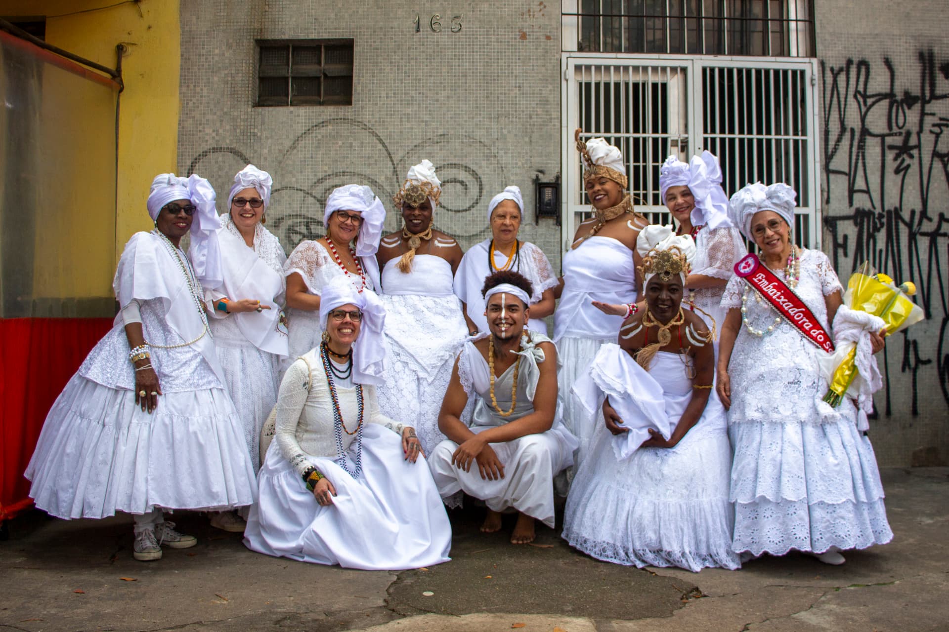 Em primeiro plano grupo de pessoas posando para a fotografia, em segundo plano, fachada de um edifício que contém pixações. As pessoas estão vestidas todas de branco com elementos vestual simbólicos de matriz africana, como, turbantes de cabeça e saias rodadas, além de algumas das pessoas estarem com pintura corporal e uma das mulheres, na ponta direita da imagem, esta vestindo uma faixa vermelha sob o peitoral escrito "Embaixadora do S{amba}" e nas mãos um buquê de flores na tonalidade amarela.