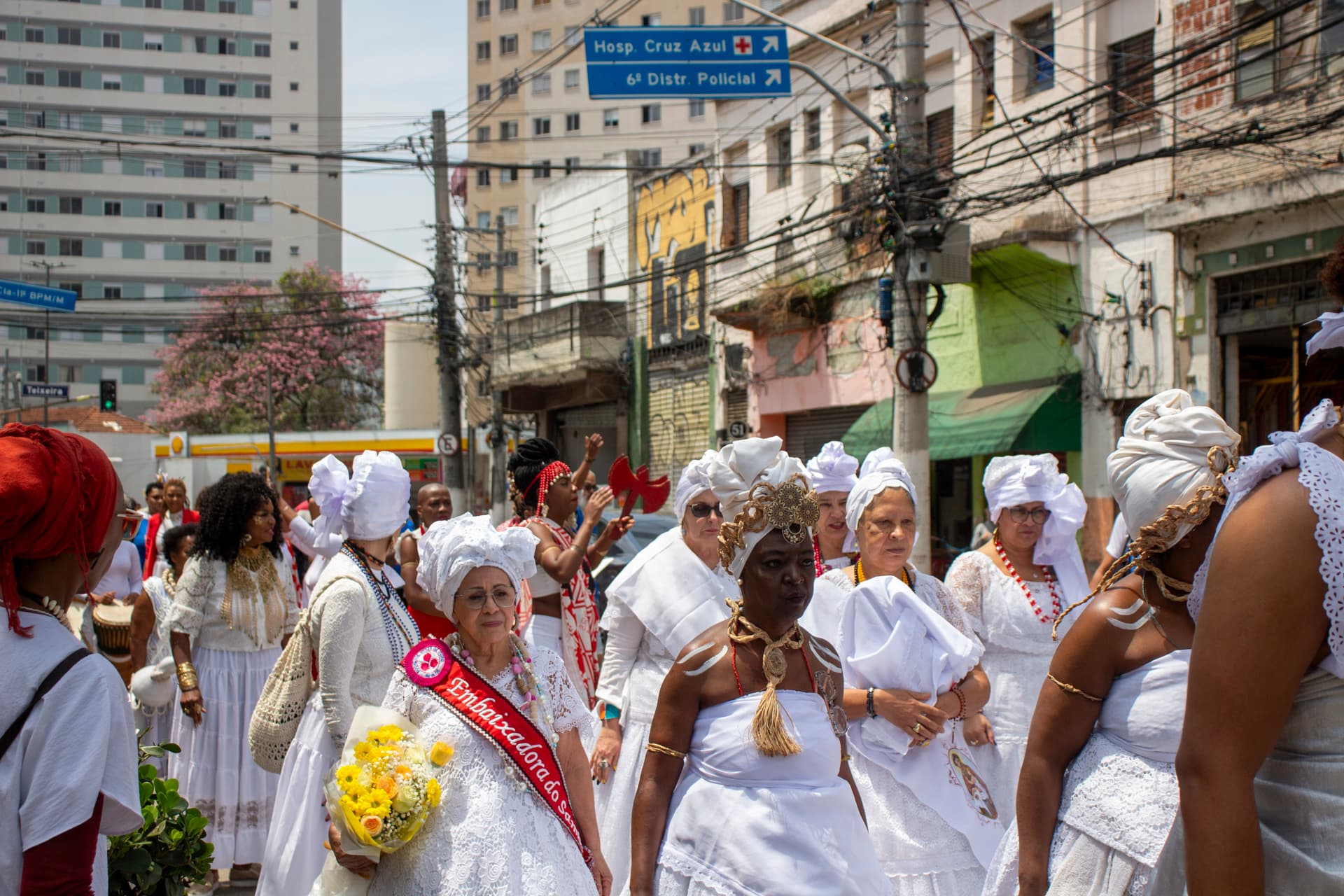 Em primeiro plano grupo de pessoas andando em cortejo na rua e olhando para o horizonte, e em segundo plano, edificações, árvore com folhagens verdes e flores rosas e placas de trânsito. As pessoas estão vestidas majoritariamente de branco com elementos vestual simbólicos de matriz africana, como, turbantes de cabeça, saias rodadas, adornos e representações de entidades, além de uma das mulheres, à esquerda da imagem, esta vestindo uma faixa vermelha sob o peitoral escrito "Embaixadora do S{amba}" e nas mãos um buquê de flores na tonalidade amarela.