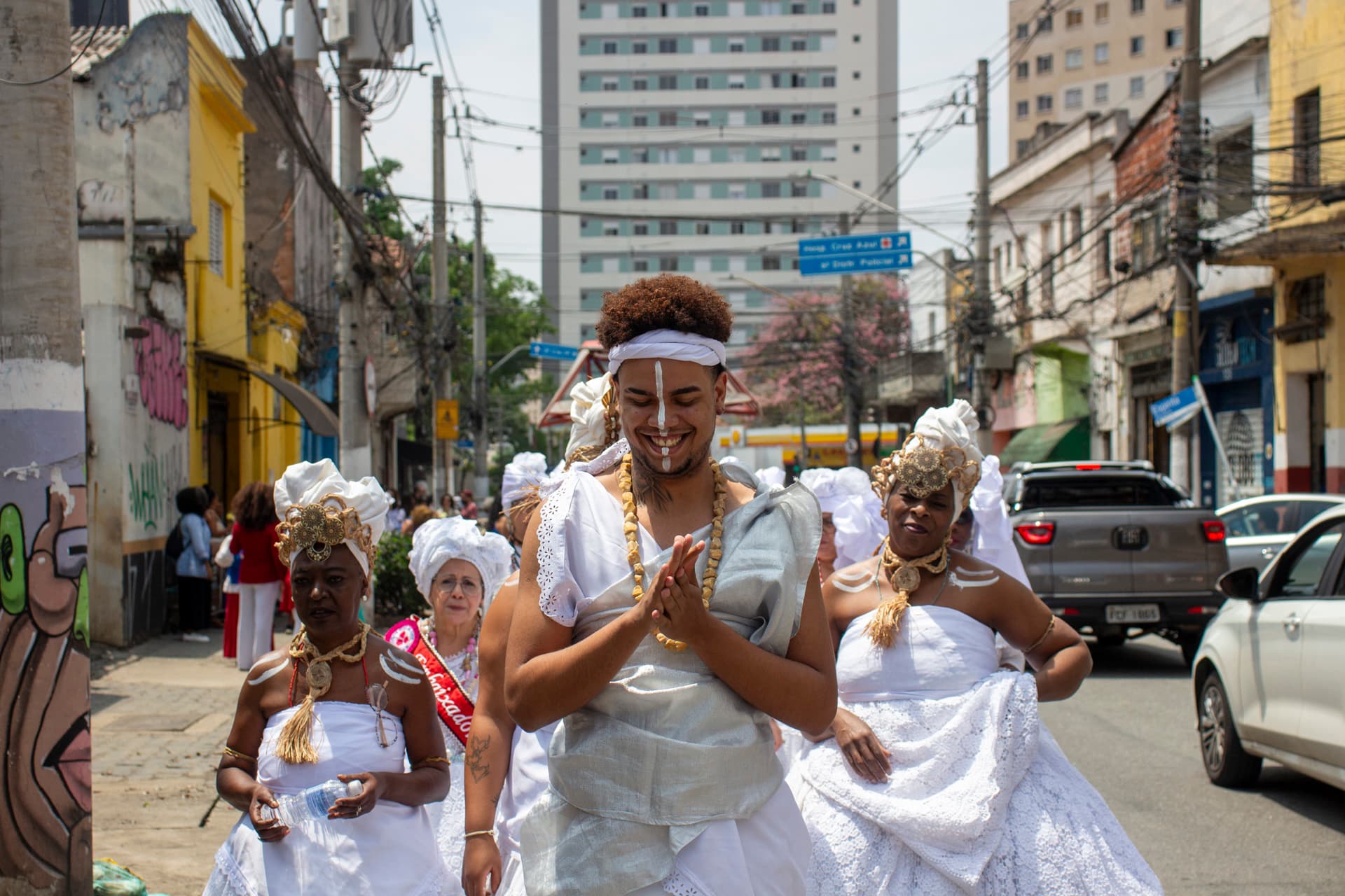 Em primeiro plano grupo de pessoas andando em cortejo na rua, tendo um jovem negro a frente de todos com expressão sorridente e mãos juntas frente ao corpo, e em segundo plano, pessoas de costas, edificações, árvores, carros e placas de trânsito. As pessoas estão vestidas majoritariamente de branco com elementos vestual simbólicos de matriz africana, como, turbantes de cabeça, saias rodadas, adornos e pinturas corporais, além de uma das mulheres, à esquerda da imagem, esta vestindo uma faixa vermelha sob o peitoral escrito "Embaixadora do S{amba}".
