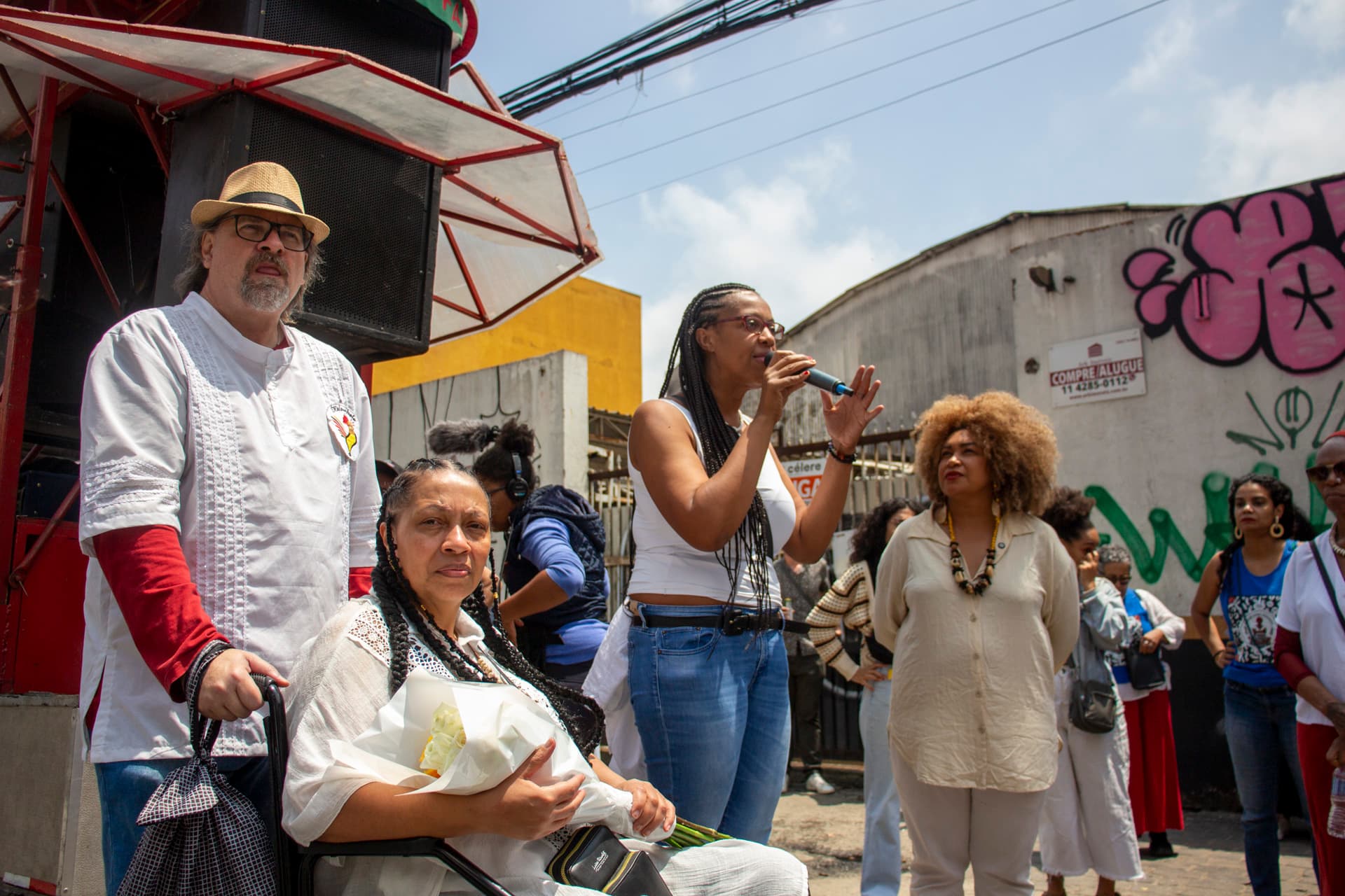 Grupo de pessoas em cortejo de rua. Em primeiro plano da esquerda para a direita homem branco com chapéu e óculos olhando para o horizonte e segurando cadeira de rodas com mulher negra de cabelo trançado segurando um buque de flores e olhando para a câmera. Em seguida mulher negra também de cabelo trançado olhando ao horizonte, falando com microfone na mão direita e com o braço esquerdo erguido com a palma da mão aberta lateralmente, ao seu lado olhando para ela outra mulher negra de cabelo crespo black. Em segundo plano, algumas pessoas acompanhando a fala ou organizando equipamentos relativos ao cortejo, um carro de som e edificações.