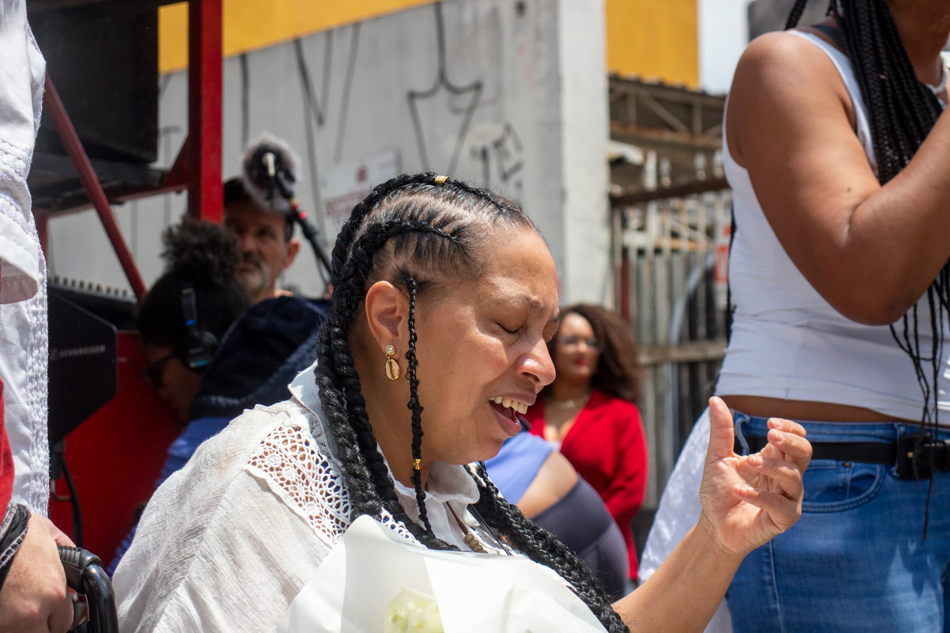 Em cortejo de rua, sentada em cadeira de rodas, mulher negra de cabelo trançado e roupa branca cantando com expressão de emoção e com a mão esquerda levantada. Em segundo plano, carro de som, pessoas e edificação.