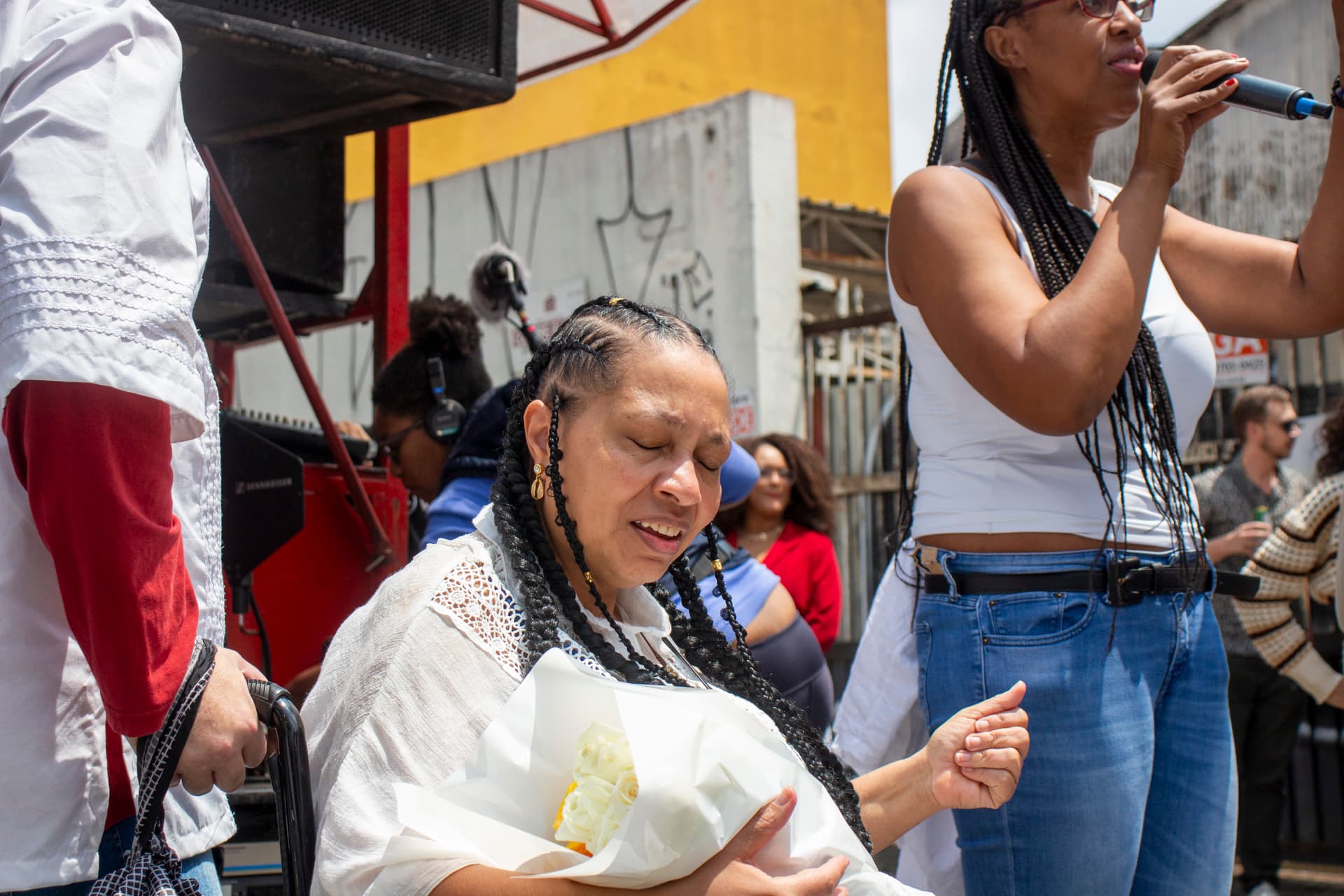 Em cortejo de rua, parte do corpo de homem branco segurando cadeira de rodas onde está sentada mulher negra de cabelo trançado e roupa branca, segurando buque de flores e cantando com expressão de emoção com a mão esquerda levantada, ao lado, outra mulher negra de cabelo trançado de blusa branca e calça jeans cantando e segurando microfone com a mão direita. Em segundo plano, carro de som, pessoas e edificação com pixação