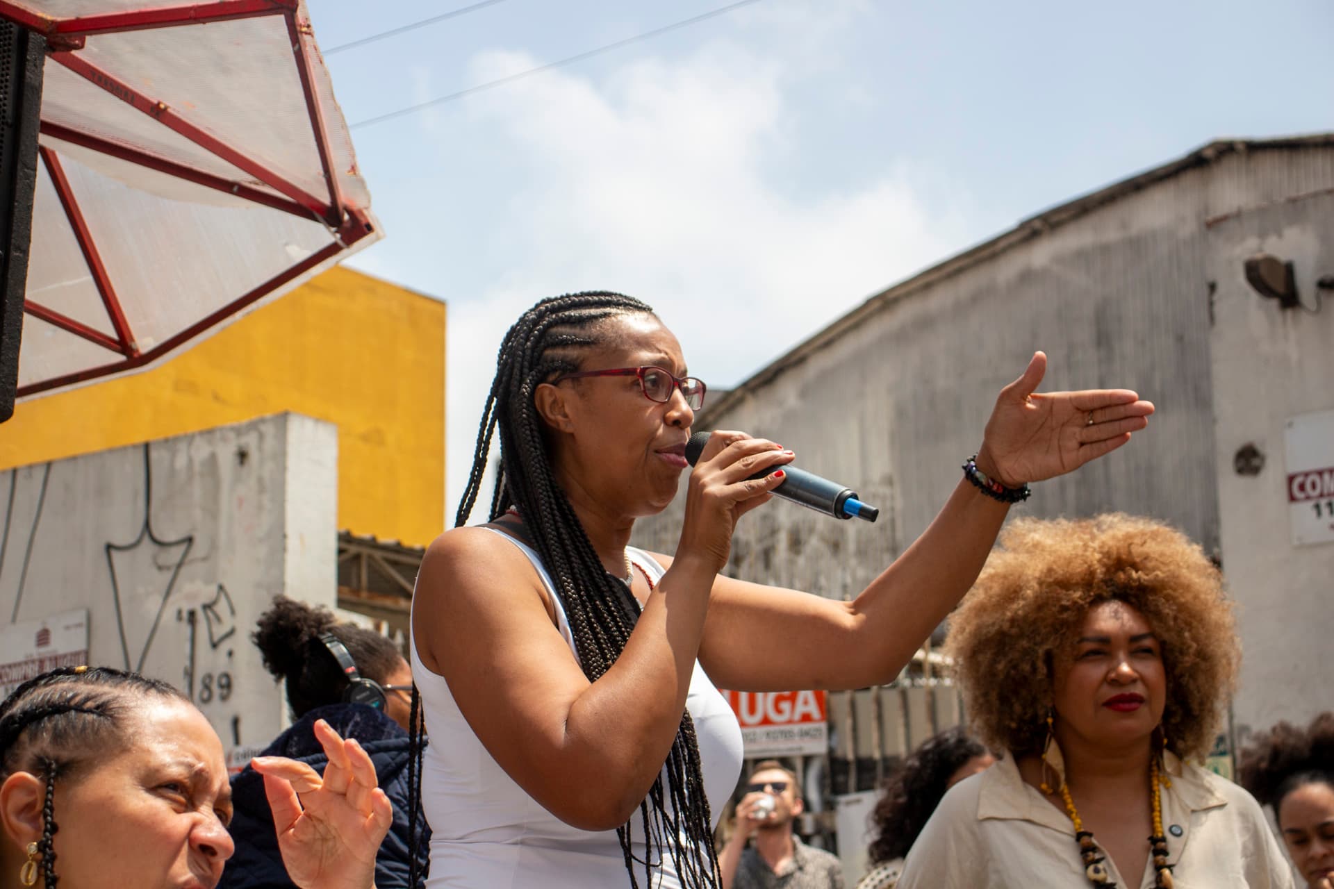 Em cortejo de rua, mulher negra de cabelo trançado com blusa branca falando com microfone na mão direita e com o braço esquerdo erguido com a palma da mão aberta lateralmente apontando para frente e olhando ao horizonte.  Ao lado esquerdo da imagem, recorte de rosto de mulher negra com a mão próximo a cabeça, ao lado direito outra mulher negra com roupa clara e cabelo crespo black, ao fundo outras pessoas e edificações.