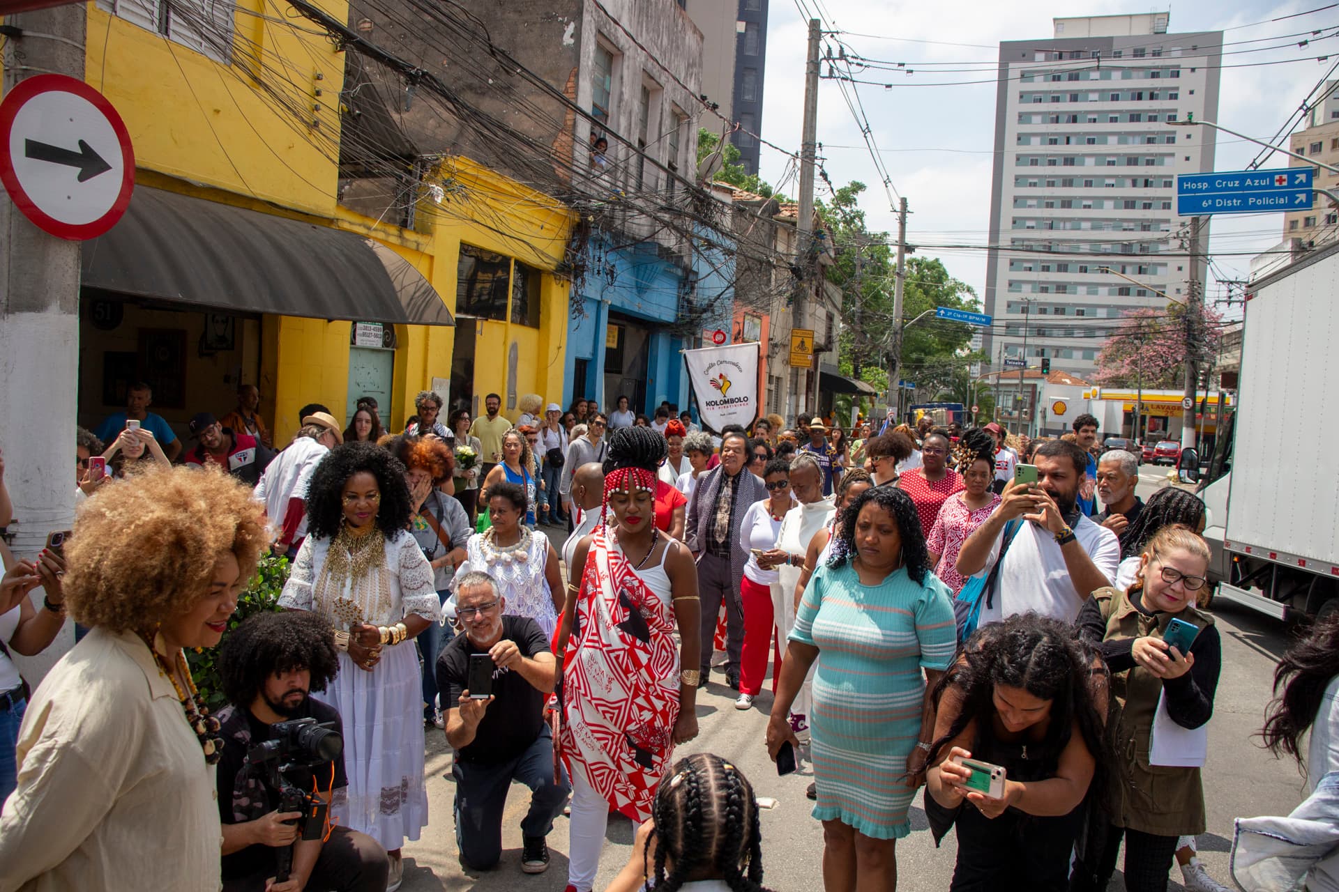 Em cortejo de rua, grupo de pessoas registrando com dispositivos tecnológicos, voltadas para uma mulher negra de costas com cabelo trançado que está falando. Ao fundo edificações, árvores, fiações de rua e placas de trânsito.