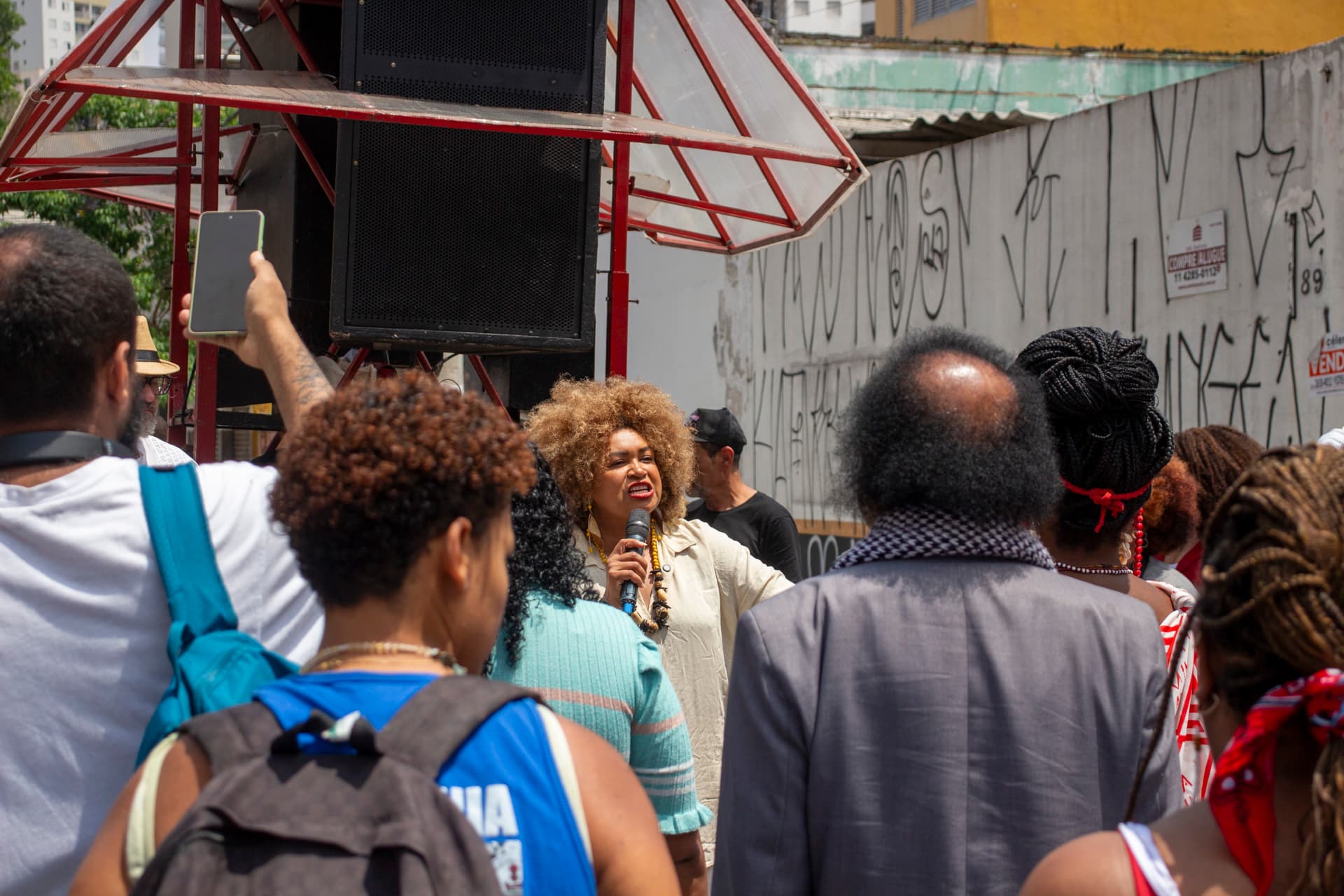 Grupo de pessoas em cortejo de rua. Em primeiro plano, pessoas de costas voltadas para mulher negra de cabelo crespo black com roupa clara e acessórios falando ao microfone. Ao fundo edificação com pixação e carro de som.