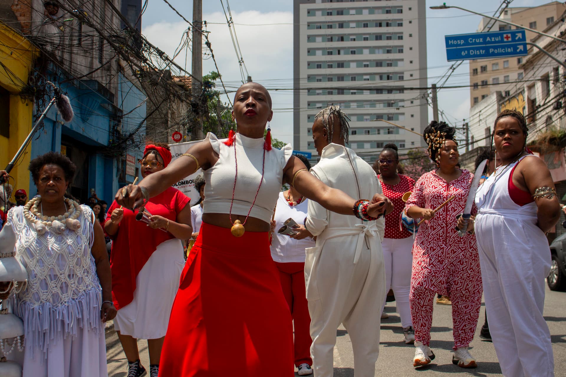 Em cortejo de rua, mulheres negras vestidas de branco e vermelho, algumas com grafismos nas roupas, acessórios e elementos simbólicos que remetem a entidades de matriz africana. As mulheres estão dançando e tocando, e, ao fundo edificações, postes com fiação elétrica e placa de trânsito indicando "Hospi.Cruz Azul" e "6º Distr. Policial".